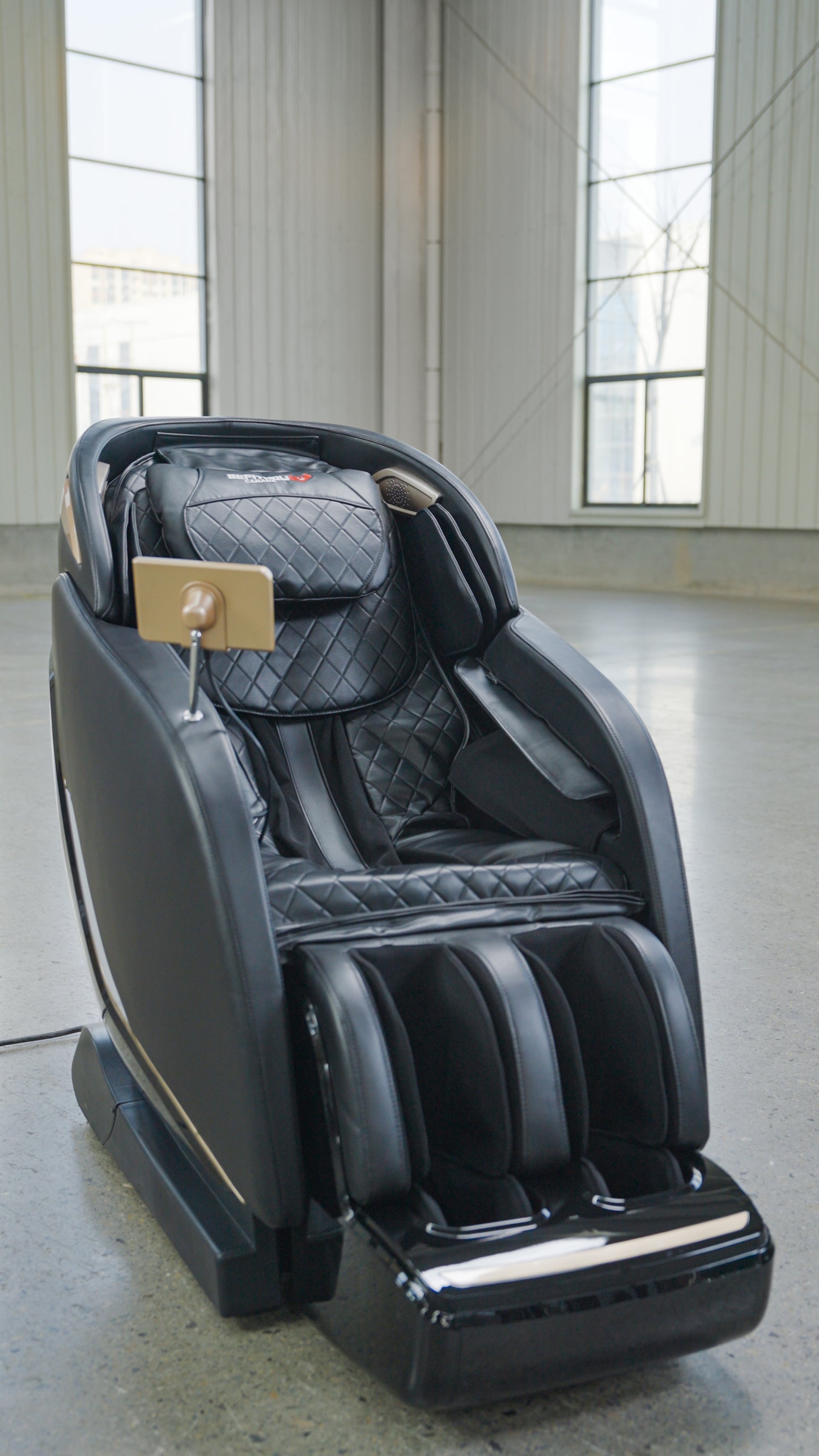 The BefitNow Canada SL Full Body Commercial Massage Chair, featuring quilted black upholstery and a touchpad control on the armrest, offers a relaxing full-body massage in spacious rooms with large windows and concrete floors.