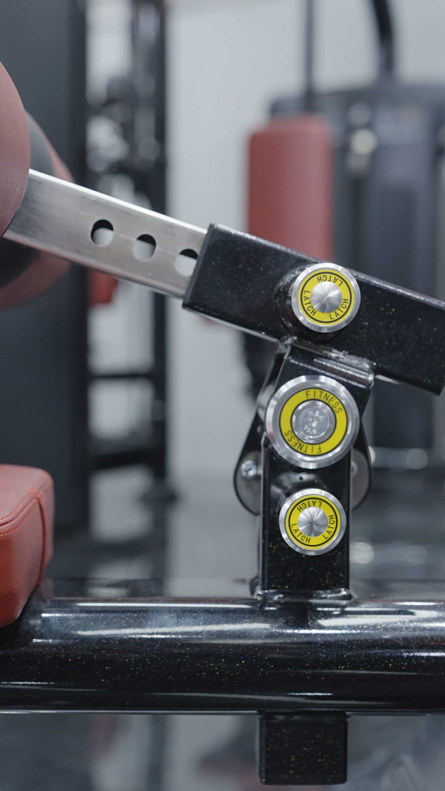 Close-up of an adjustable weight bench with red padding and yellow knobs in a gym, with a BefitNow Canada Iso Lateral Lat Pulldown/Low Row machine visible among blurred commercial-grade equipment in the background.