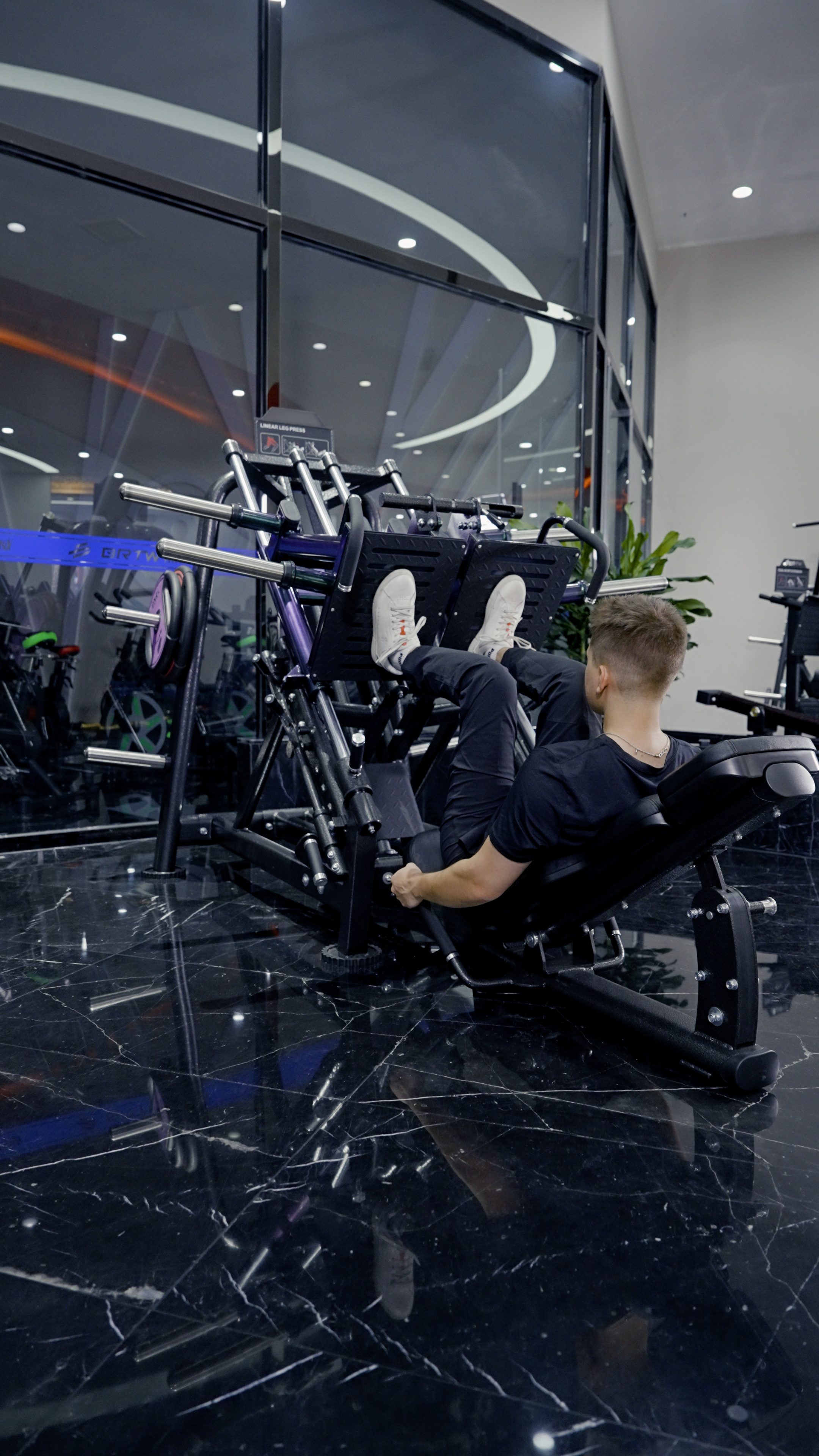 A man in black athletic wear trains his lower body at night on the BefitNow Canada Quadzilla Commercial ISO Leg Press in a modern gym with black marble floors and large glass windows.