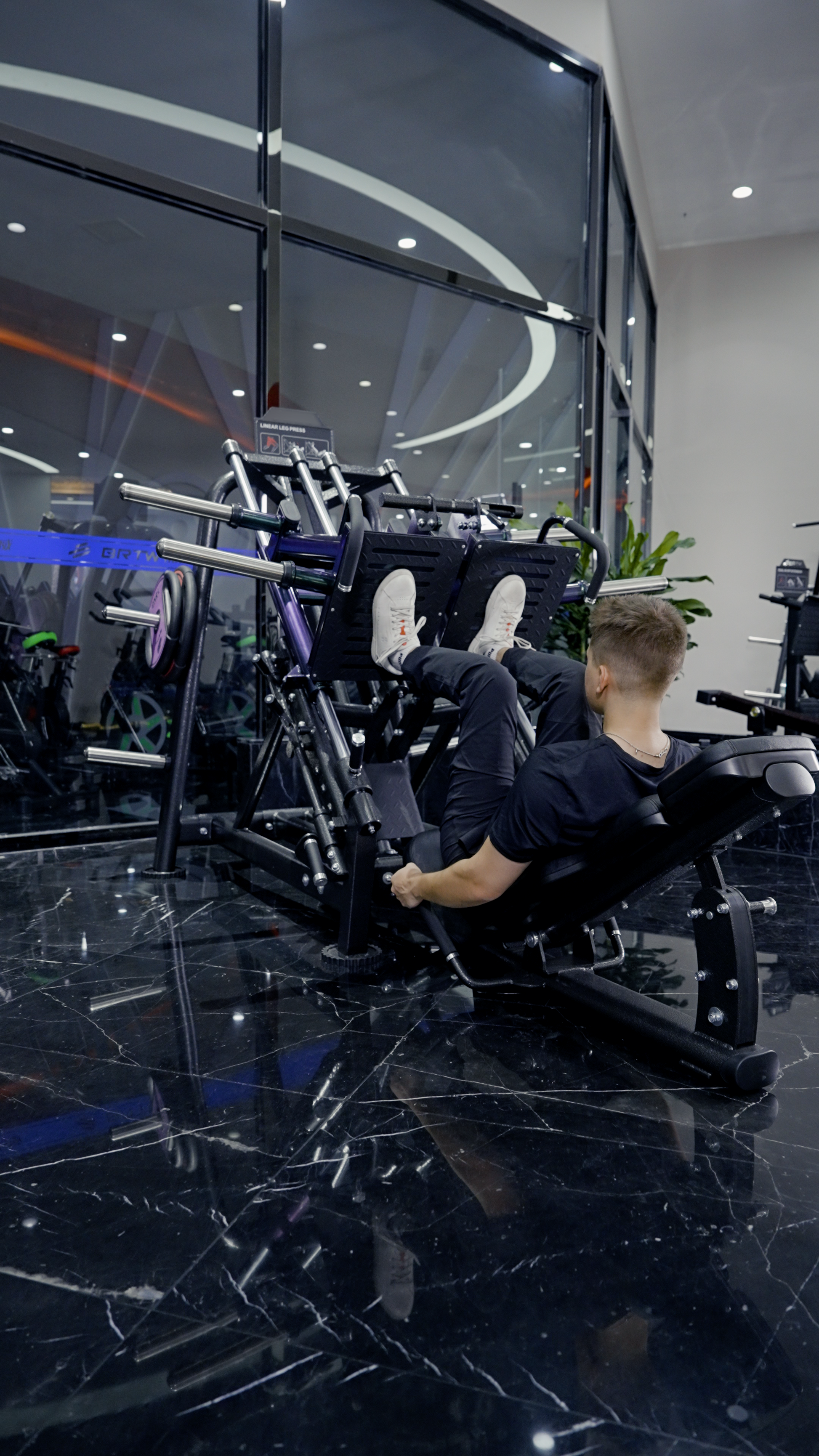 A man in black athletic wear trains his lower body at night on the BefitNow Canada Quadzilla Commercial ISO Leg Press in a modern gym with black marble floors and large glass windows.