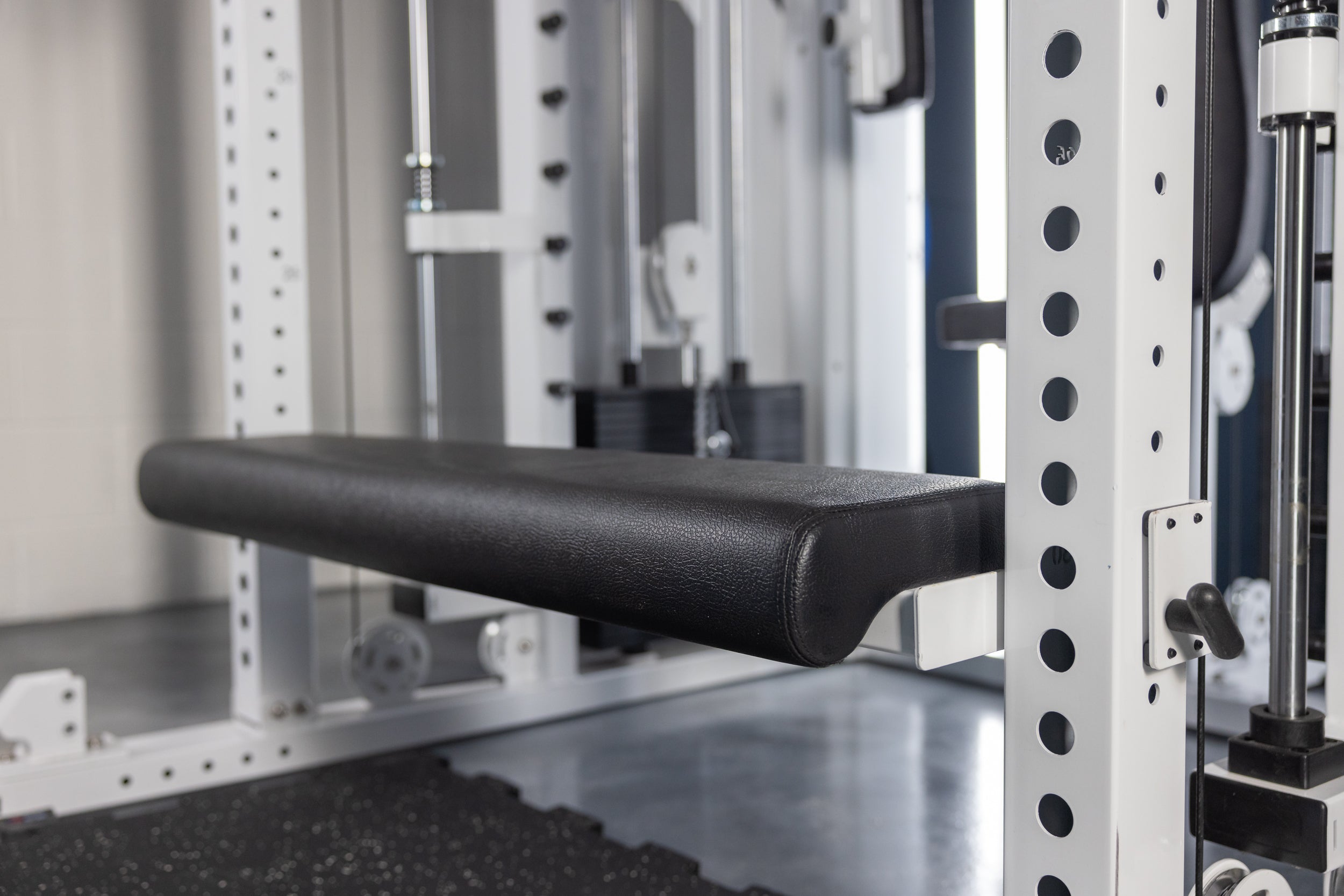 Close-up of a black padded bench inside The Kahuna All in One Power Rack by BefitNow Canada, featuring a commercial steel frame with multiple adjustment holes, weight stacks, and integrated trainer equipment in the gym.