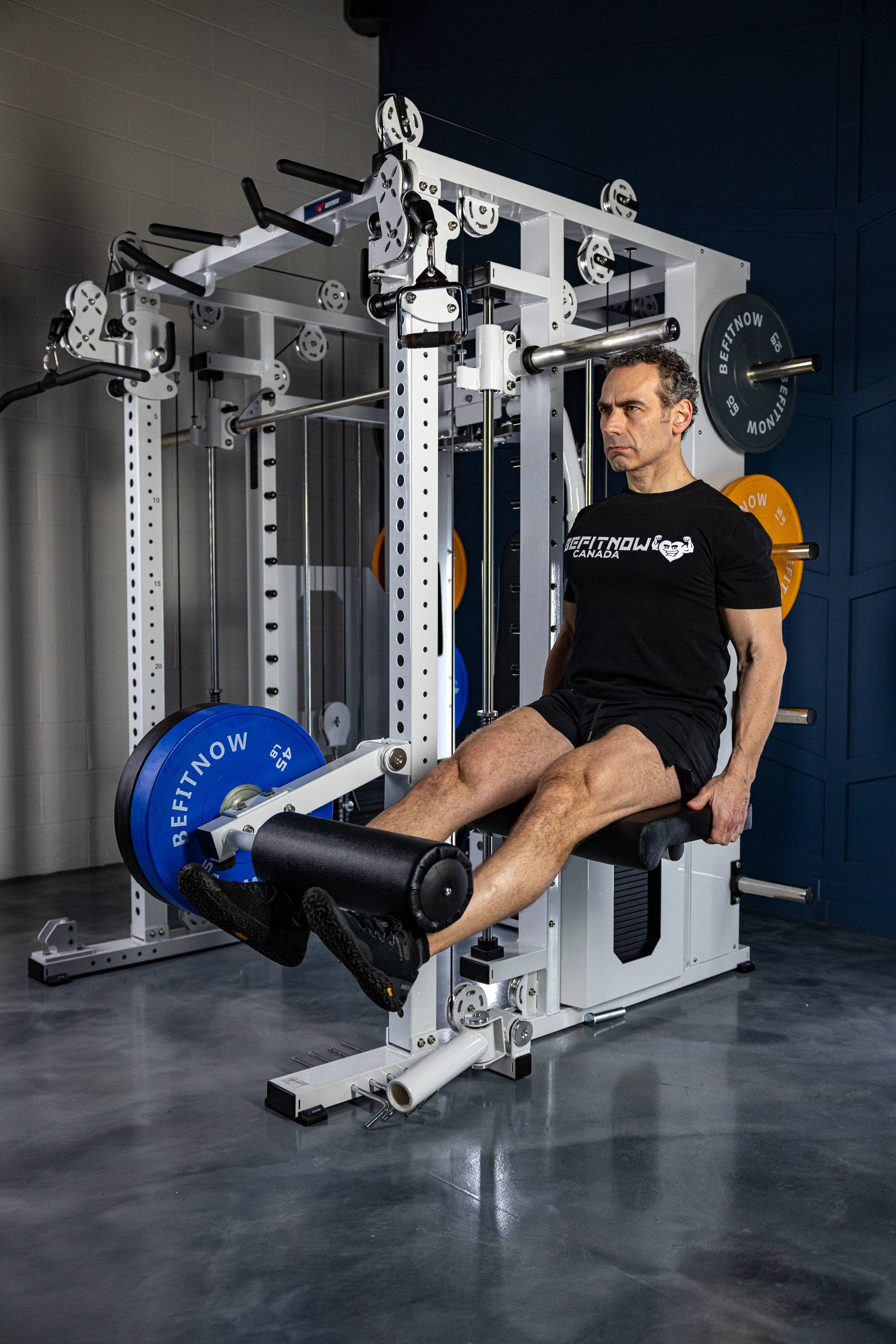 A man wearing a black T-shirt and shorts performs a leg extension on the BefitNow Canada Kahuna All in One Power Rack, surrounded by weight plates, fitness equipment, and sturdy commercial steel frames in the gym.