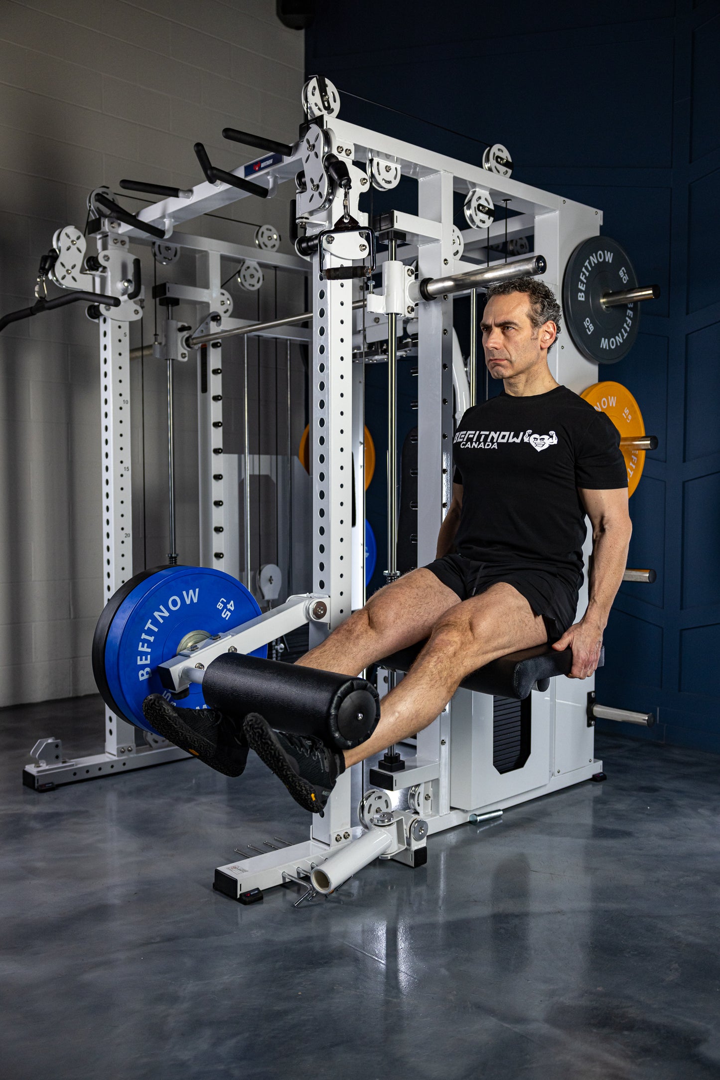 A man wearing a black T-shirt and shorts performs a leg extension on the BefitNow Canada Kahuna All in One Power Rack, surrounded by weight plates, fitness equipment, and sturdy commercial steel frames in the gym.