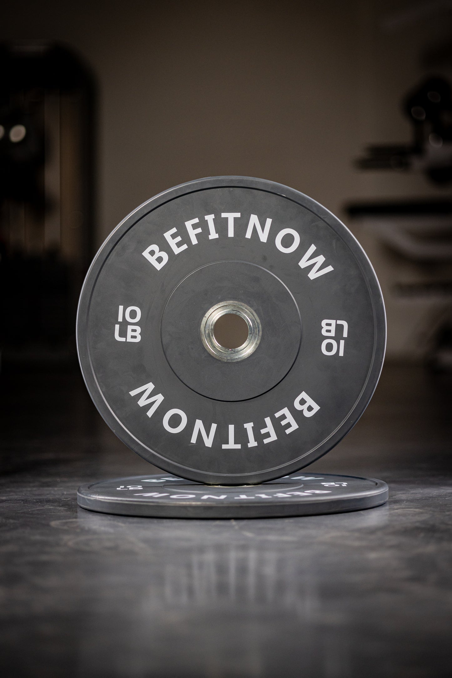 A close-up of a black 10 lb Coloured Bumper Plate by BefitNow Canada, labeled BEFITNOW, stands upright on a gym floor with another plate flat underneath—ideal for Olympic weightlifting and strength training.