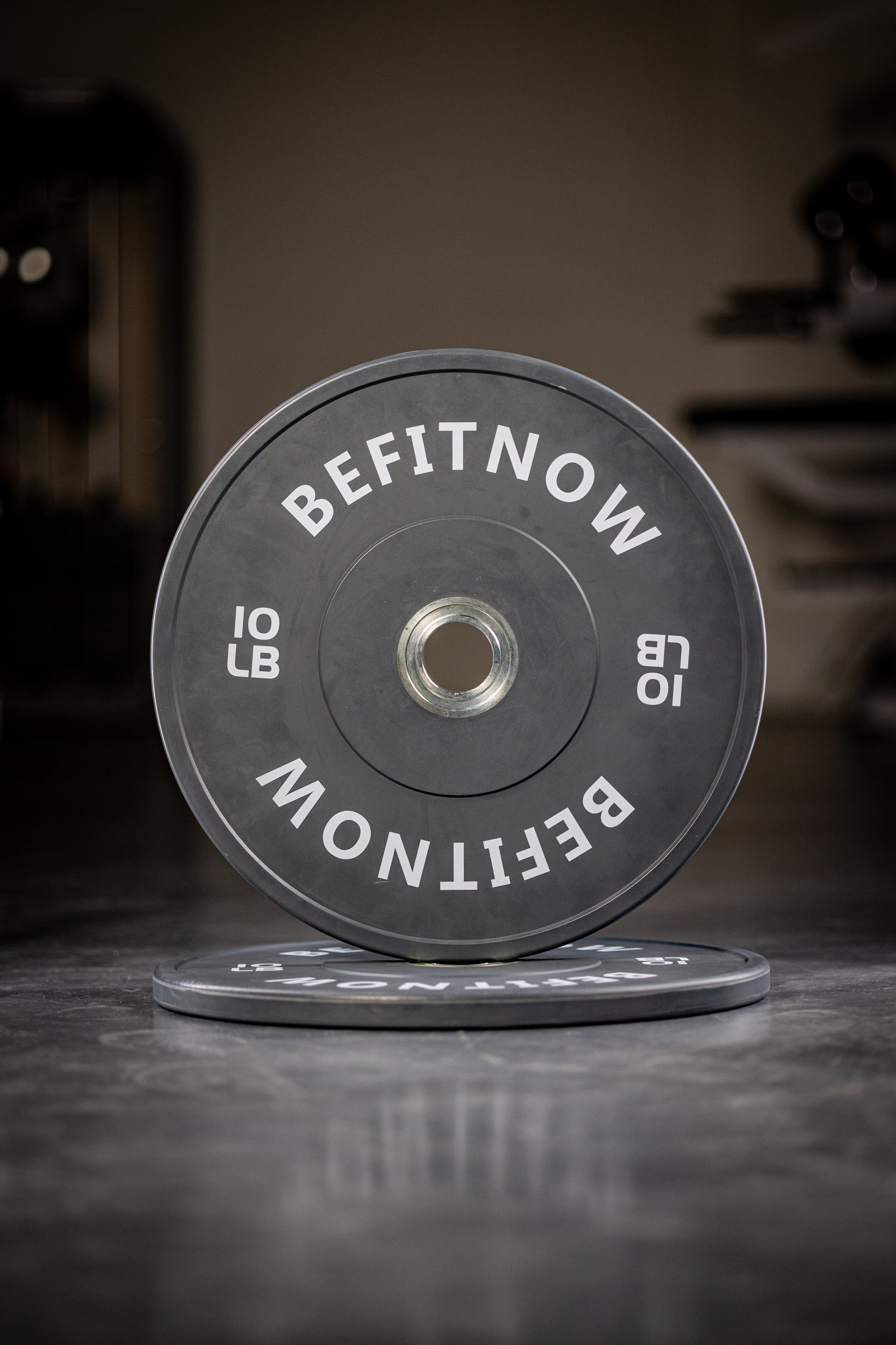 A close-up of a black 10 lb Coloured Bumper Plate by BefitNow Canada, labeled BEFITNOW, stands upright on a gym floor with another plate flat underneath—ideal for Olympic weightlifting and strength training.