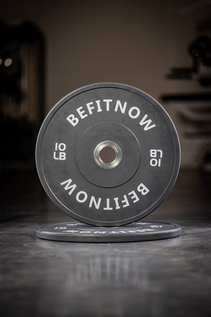A close-up of a black 10 lb Coloured Bumper Plate by BefitNow Canada, labeled BEFITNOW, stands upright on a gym floor with another plate flat underneath—ideal for Olympic weightlifting and strength training.