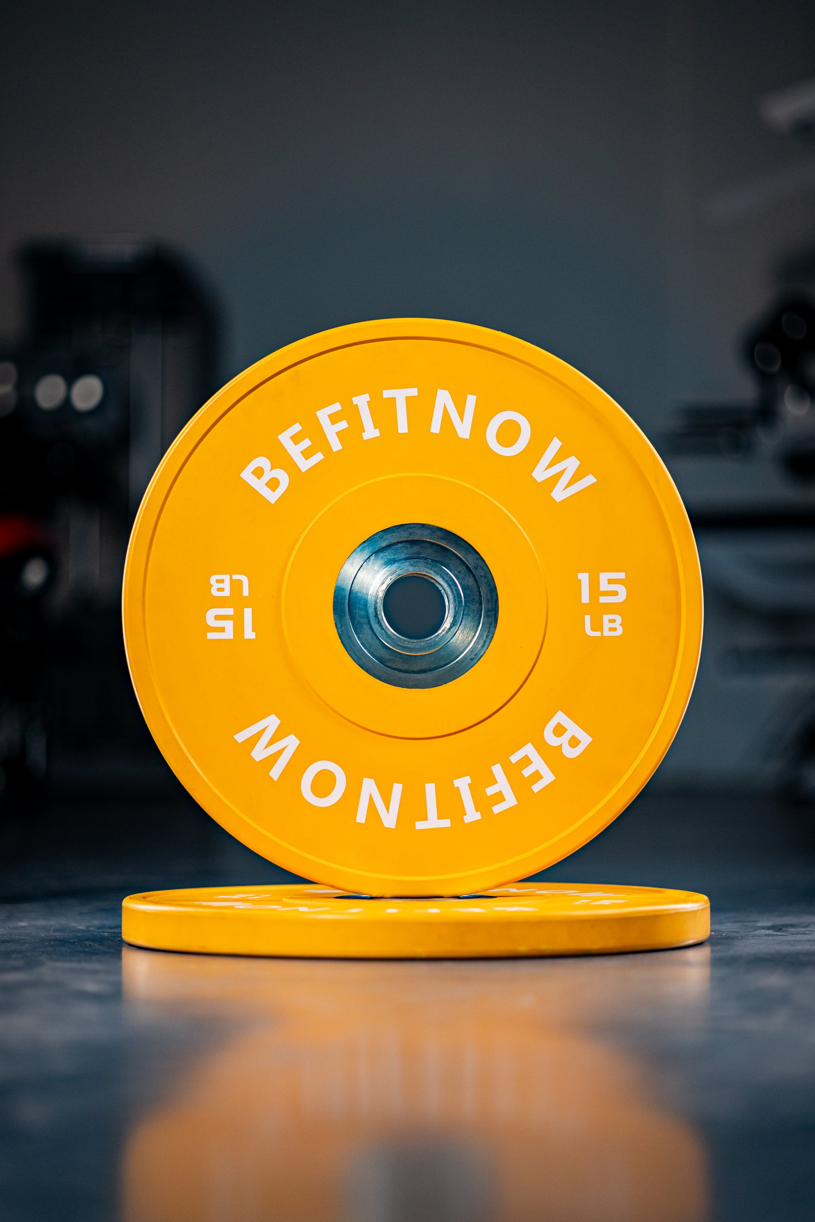 A bright yellow 15 lb Coloured Bumper Plate by BefitNow Canada stands upright, leaning against another identical plate on the gym floor—ideal for Olympic weightlifting and excellent shock absorption.