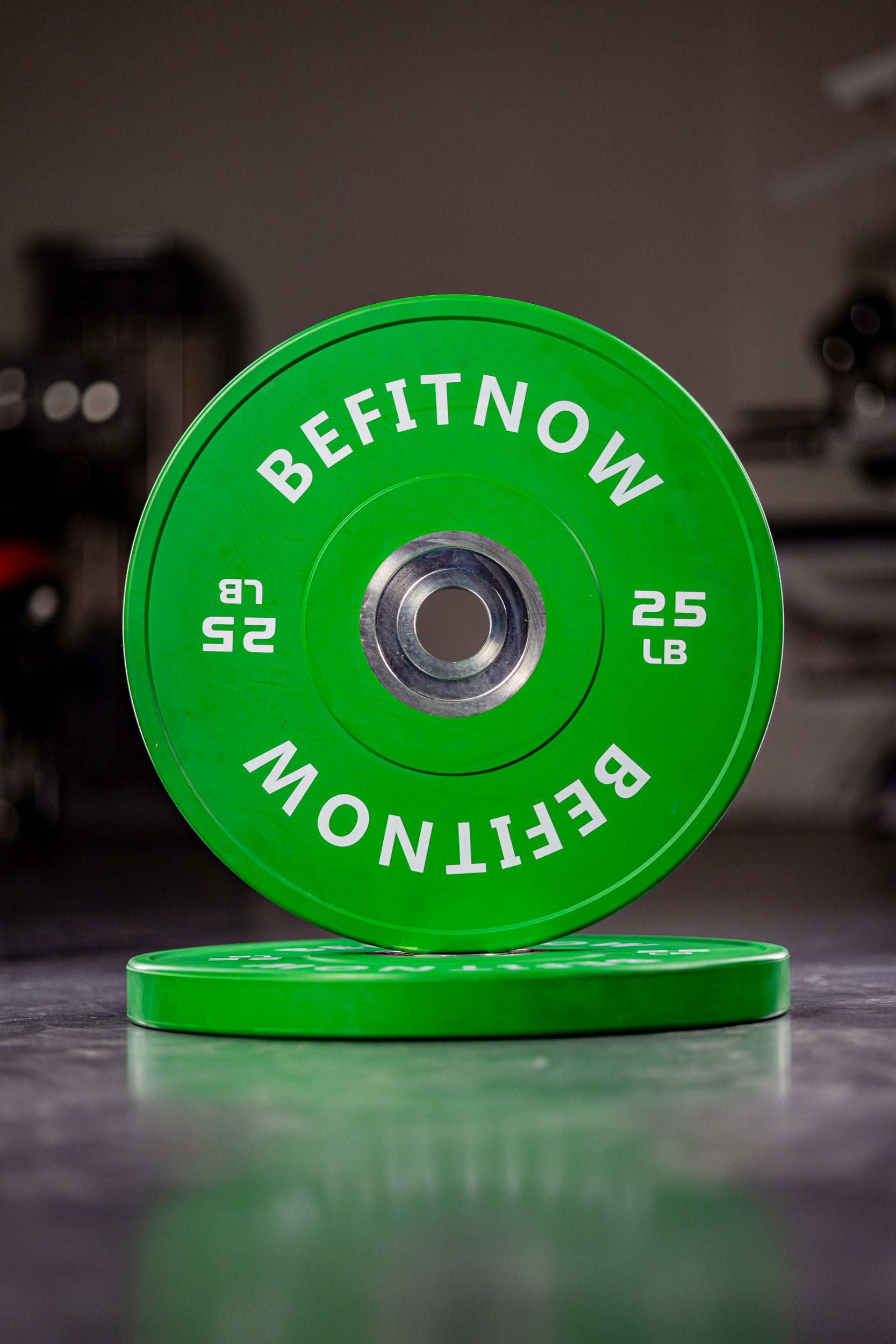 A green 25lb Coloured Bumper Plate from BefitNow Canada stands upright atop another on a gym floor. Designed for shock absorption, theyre perfect for Olympic lifts, with blurred gym equipment visible in the background.