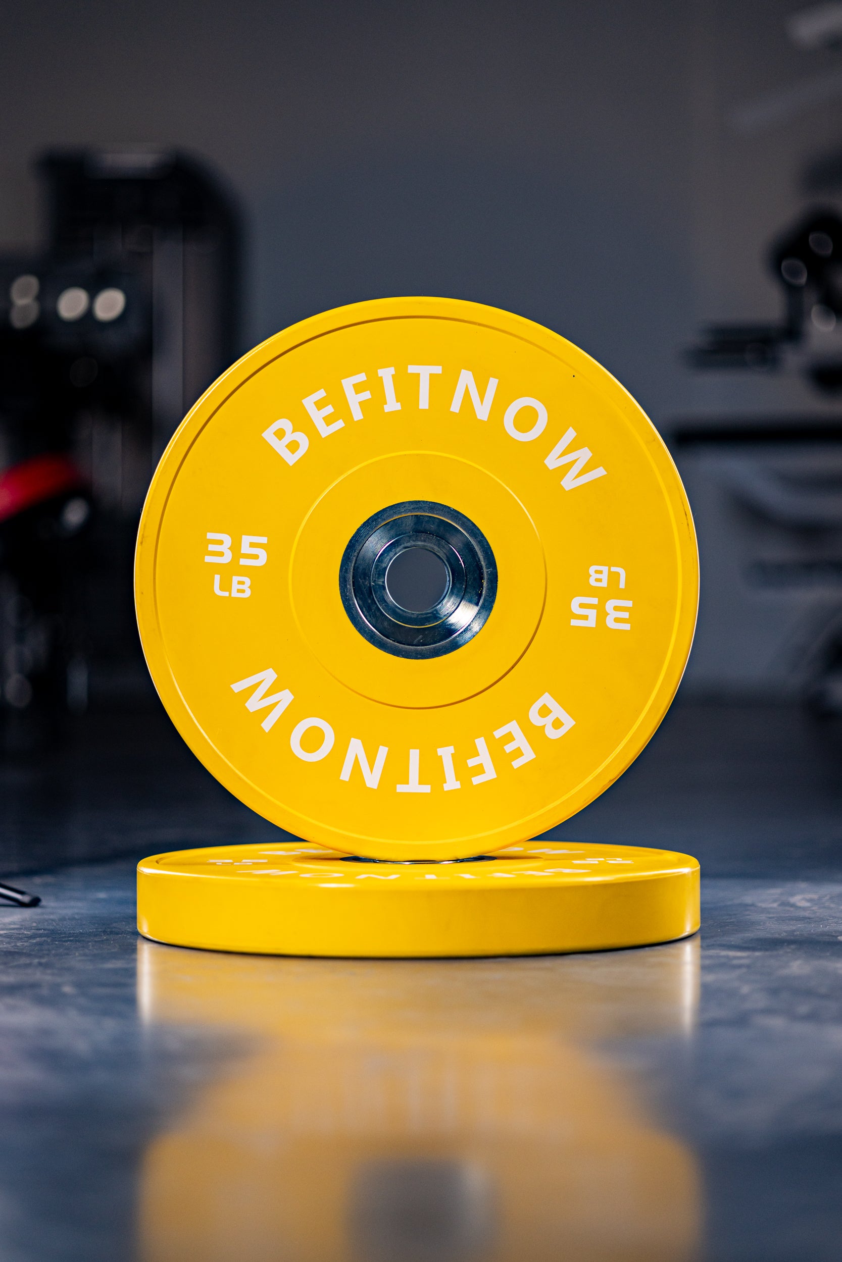 Two bright yellow Coloured Bumper Plates from BefitNow Canada, each 35 lb and labeled BEFITNOW, are displayed—one upright and one flat on the gym floor. Designed for Olympic lifting, they offer great shock absorption against a blurred gym background.