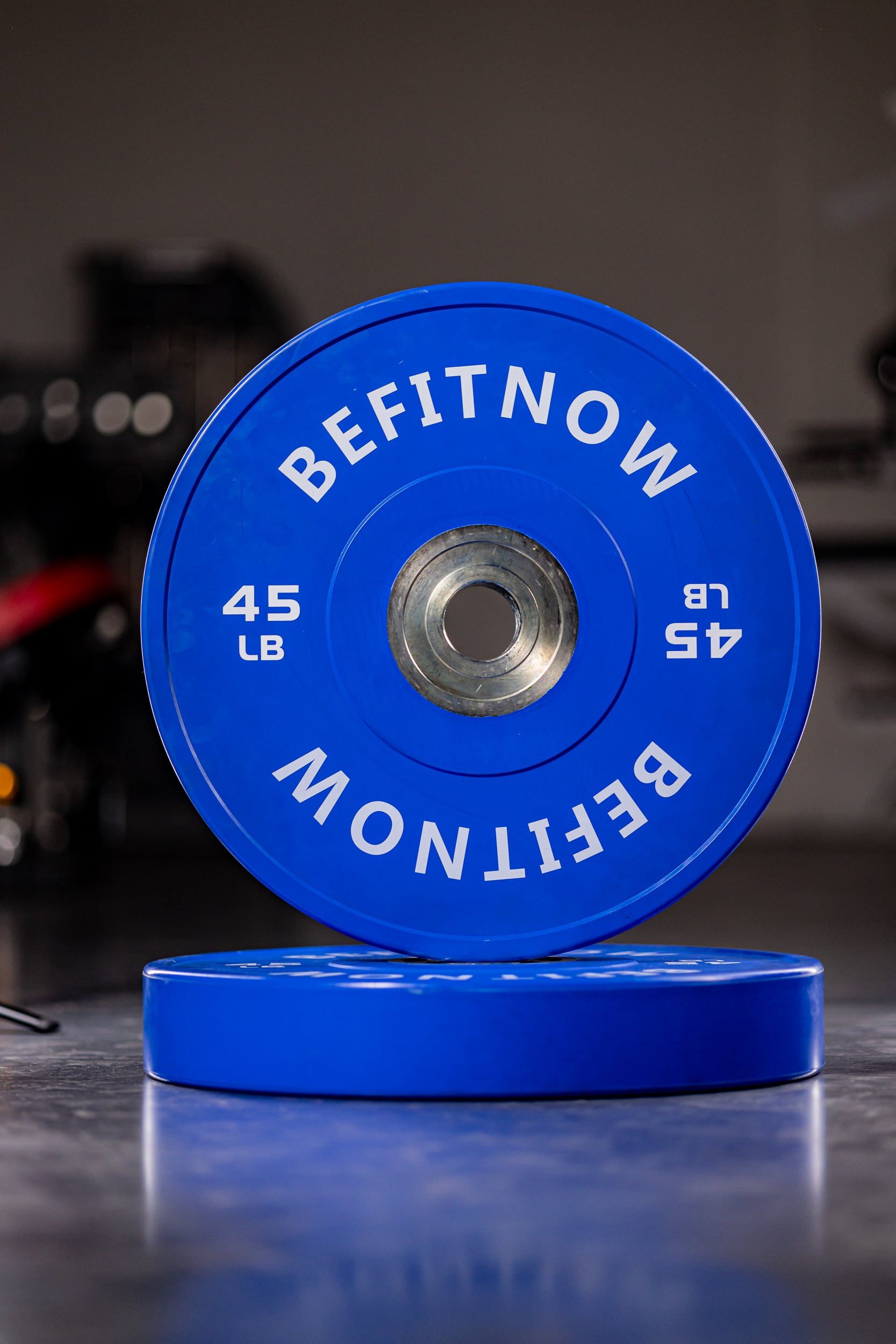 Two blue 45-lb BefitNow Canada Coloured Bumper Plates, one standing on another flat, both with BEFITNOW in white lettering. Ideal for Olympic weightlifting. The background is blurred.