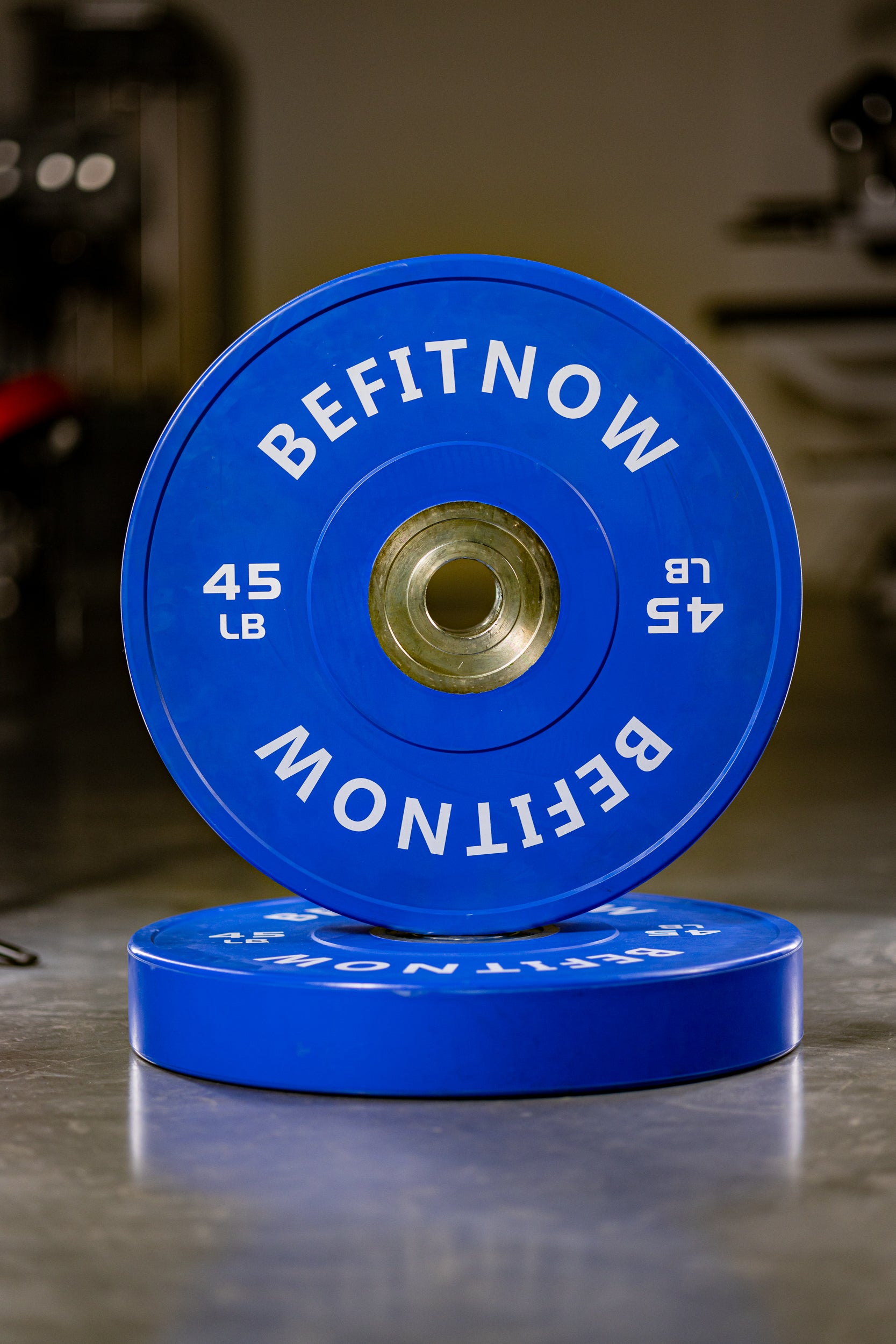A blue 45-pound Coloured Bumper Plate from BefitNow Canada stands upright on a matching plate in a gym, perfect for Olympic weightlifting, with blurred fitness equipment visible in the background.