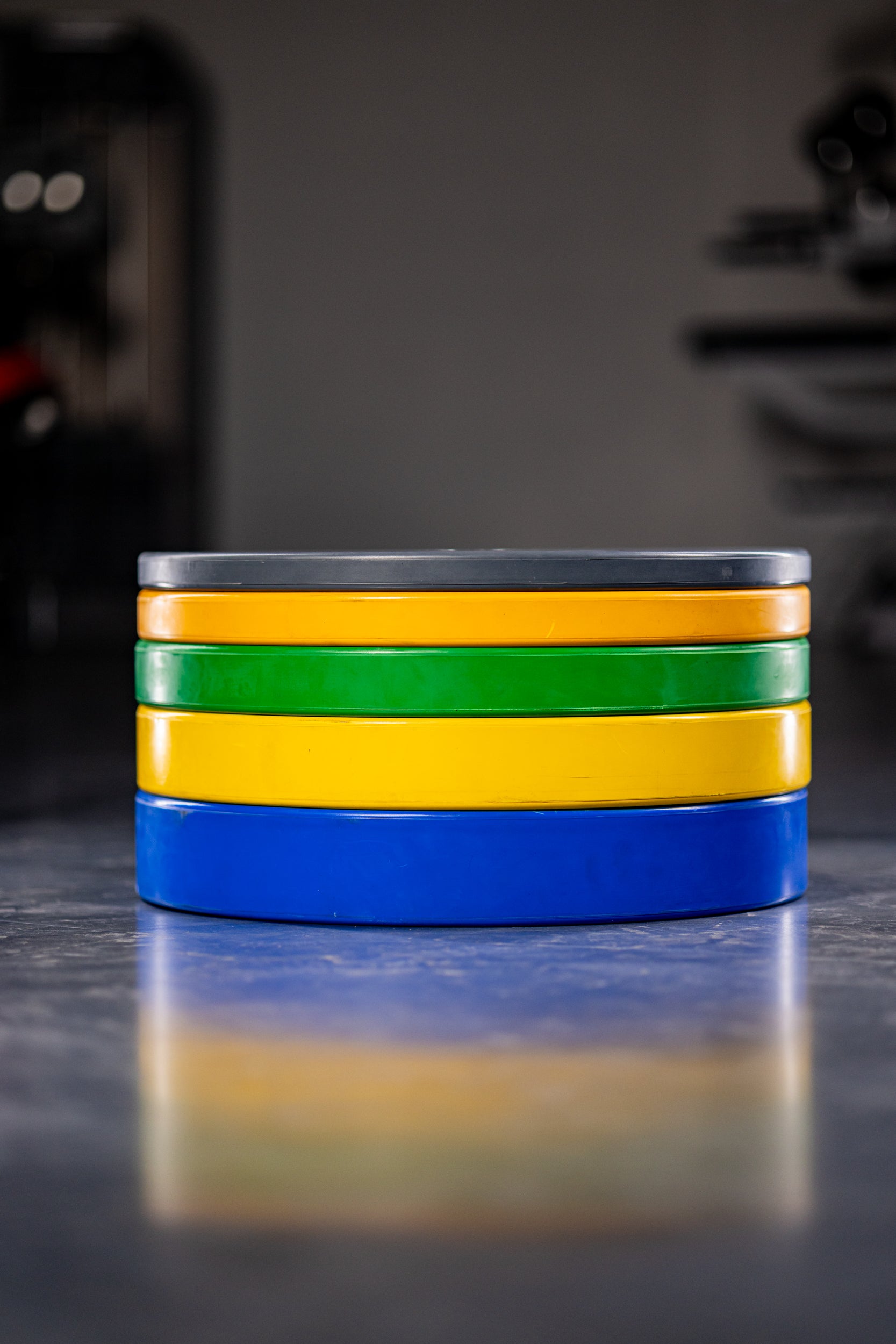 Four stacked Coloured Bumper Plates by BefitNow Canada in gray, yellow, green, and blue rest on a shiny floor with a blurred gym background. Ideal for Olympic weightlifting, these plates offer excellent shock absorption.