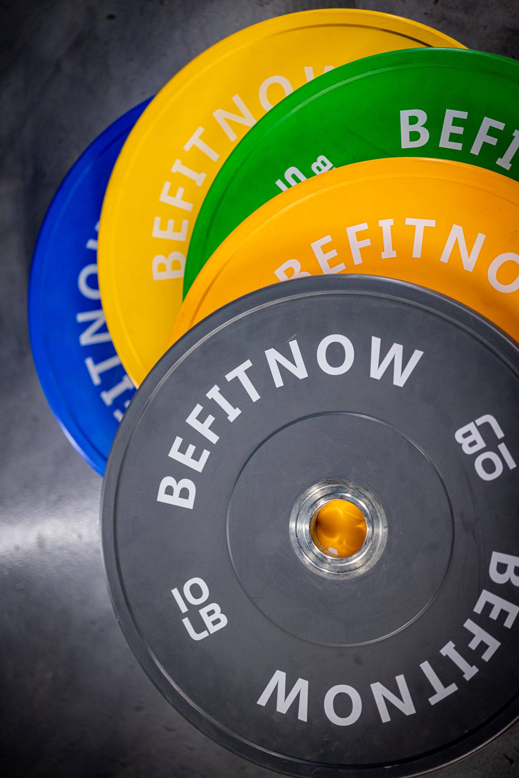 A close-up of BefitNow Canada Coloured Bumper Plates in blue, yellow, green, and black overlapping on a gym floor. Ideal for Olympic weightlifting with shock absorption to protect weights and floors. The front black plate displays 10 LB in white text.