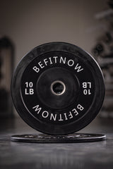 A close-up of a black 10-pound BefitNow Canada Bumper Plate labeled BEFITNOW stands upright on the gym floor, known for excellent shock absorption and ideal for Olympic weightlifting, with another plate lying flat in the background amidst blurred gym equipment.