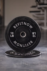 A 10-pound black Bumper Plate from BefitNow Canada, sporting its logo in white, leans against an identical plate on the gym floor. The blurred background suggests Olympic weightlifting gear, emphasizing the plates shock absorption advantage.