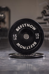 Close-up of two black 15 lb Bumper Plates by BefitNow Canada, featuring BEFITNOW text. One stands upright while the other lies flat, highlighting their shock absorption. Blurred gym equipment is in the background.