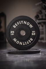 Two black BefitNow Canada Bumper Plates marked 25 LB are stacked on a gym floor. The front plate is upright, ideal for Olympic weightlifting, with blurred gym equipment in the background.