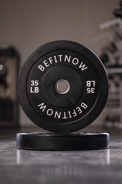 A close-up of a 35-pound BefitNow Canada black Bumper Plate, marked with BEFITNOW in white, stands upright with its twin flat behind it. Designed for Olympic weightlifting, these plates offer excellent shock absorption against blurred gym equipment.