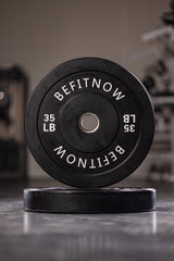 A close-up of a 35-pound BefitNow Canada black Bumper Plate, marked with BEFITNOW in white, stands upright with its twin flat behind it. Designed for Olympic weightlifting, these plates offer excellent shock absorption against blurred gym equipment.