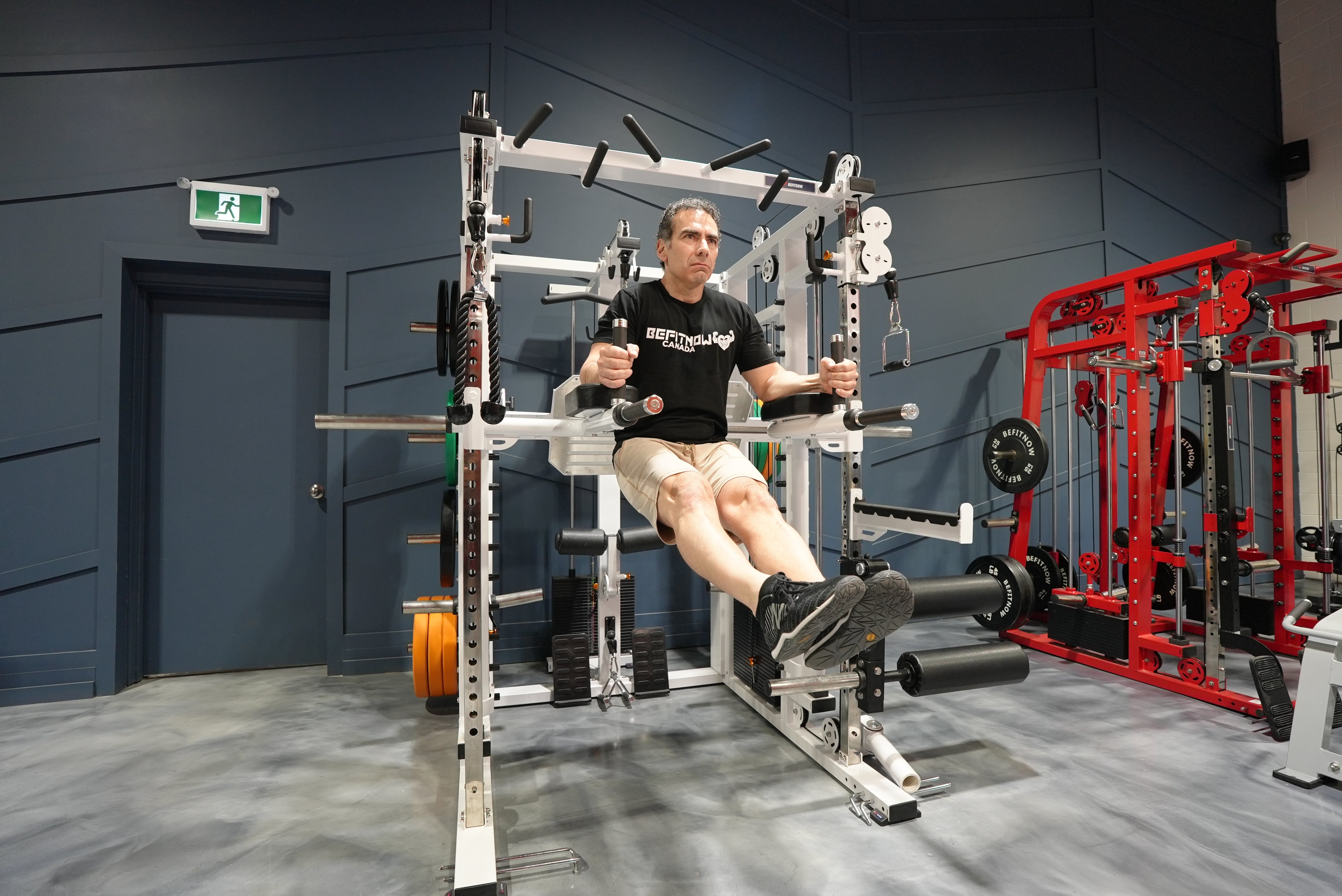 A man in a black T-shirt and beige shorts performs a hanging leg raise on The Relentless Trainer Smith Machine by BefitNow Canada in a modern gym with diverse weightlifting equipment.