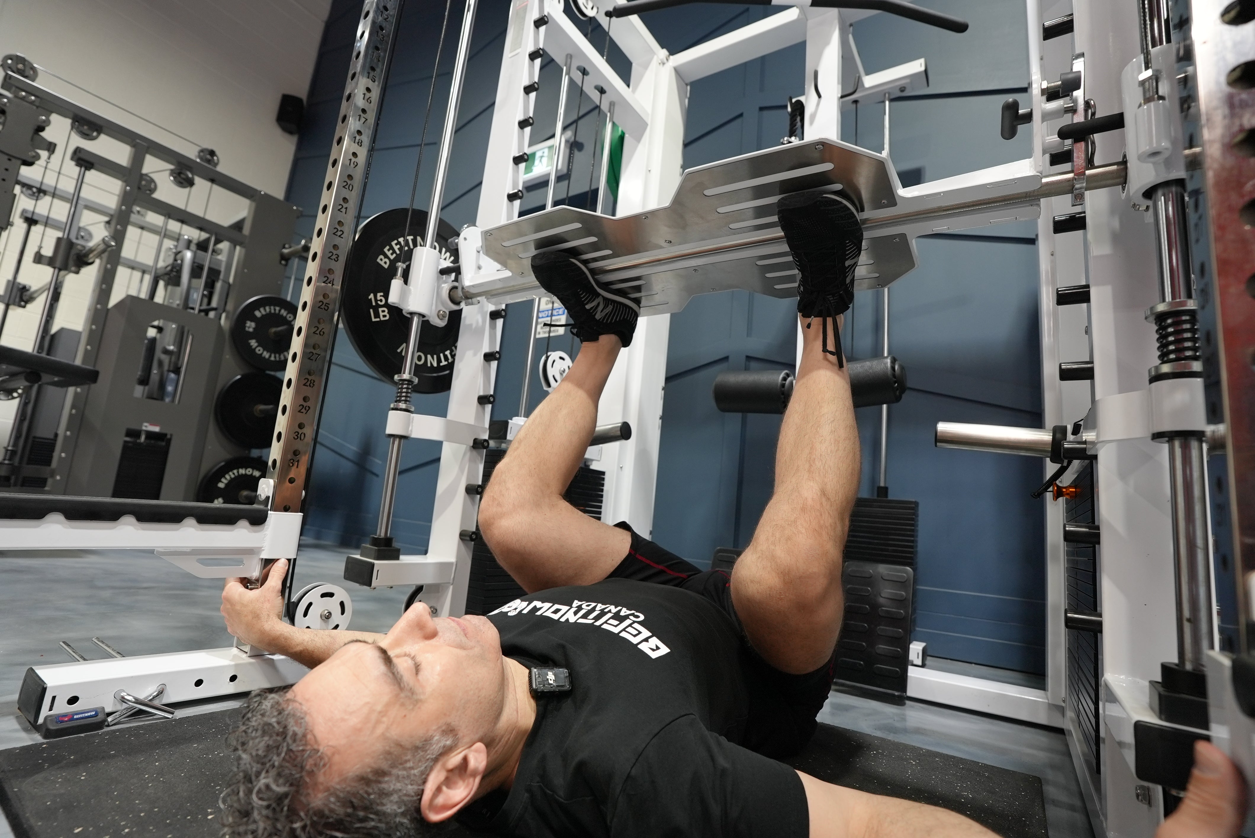 A man lies on his back in a gym using the BefitNow Canada Relentless Trainer Smith Machine to do a leg press, pushing the weighted platform up with his feet and gripping the side of the all-in-one trainer for support.