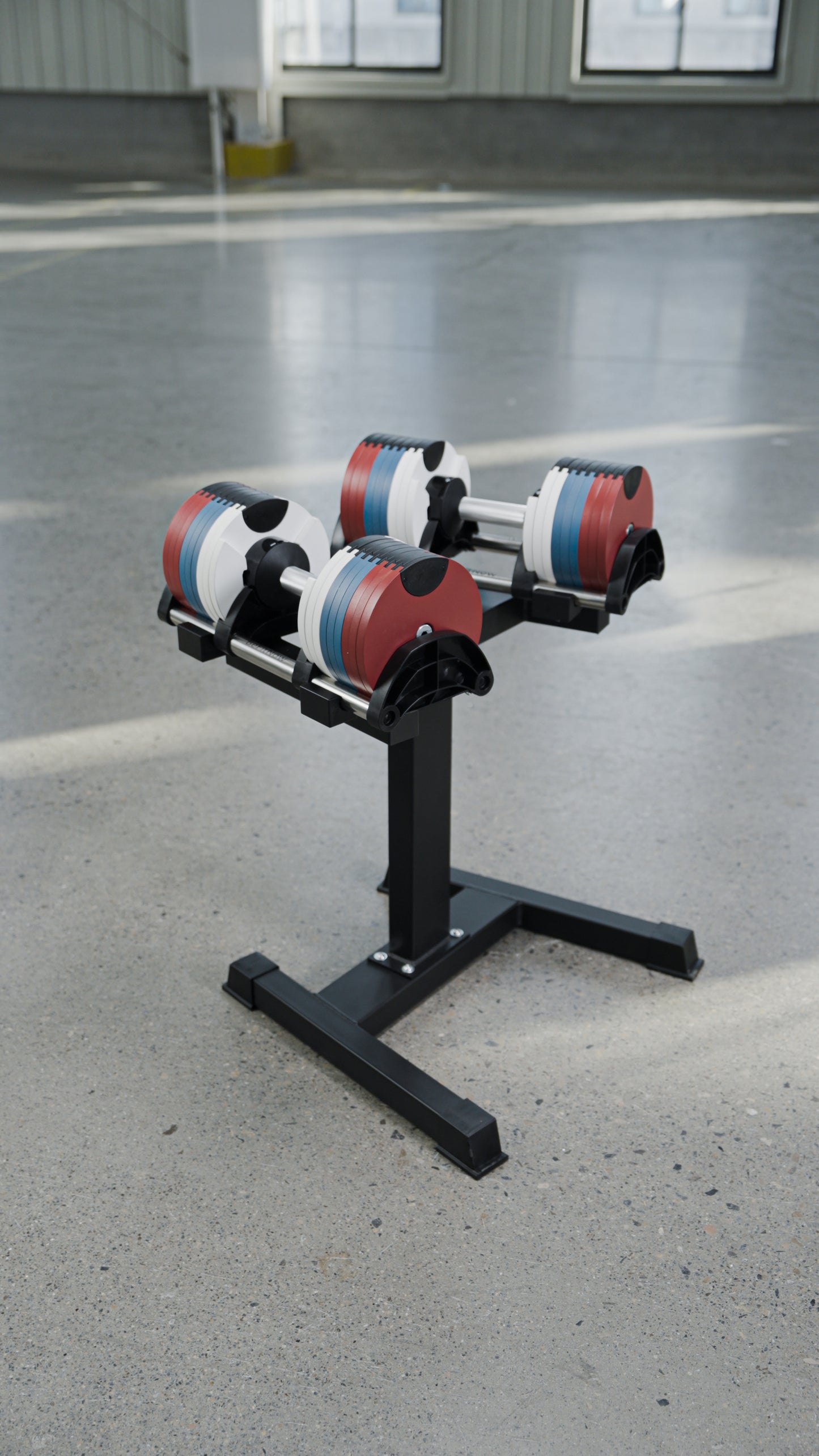 Two BefitNow Canada 80lbs Adjustable Cast Iron Dumbbells, featuring red, white, and blue weight plates, rest on a sleek black metal stand set atop a smooth light gray concrete floor in a spacious, well-lit room—an ideal choice for anyone seeking to upgrade their home gym with stylish and space-saving dumbbells.