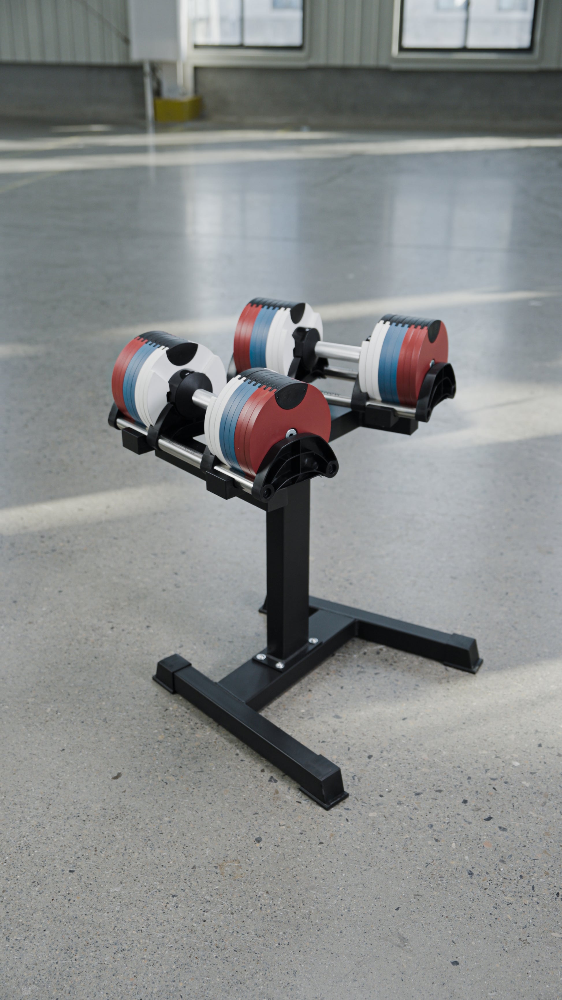 Two BefitNow Canada 80lbs Adjustable Cast Iron Dumbbells, featuring red, white, and blue weight plates, rest on a sleek black metal stand set atop a smooth light gray concrete floor in a spacious, well-lit room—an ideal choice for anyone seeking to upgrade their home gym with stylish and space-saving dumbbells.