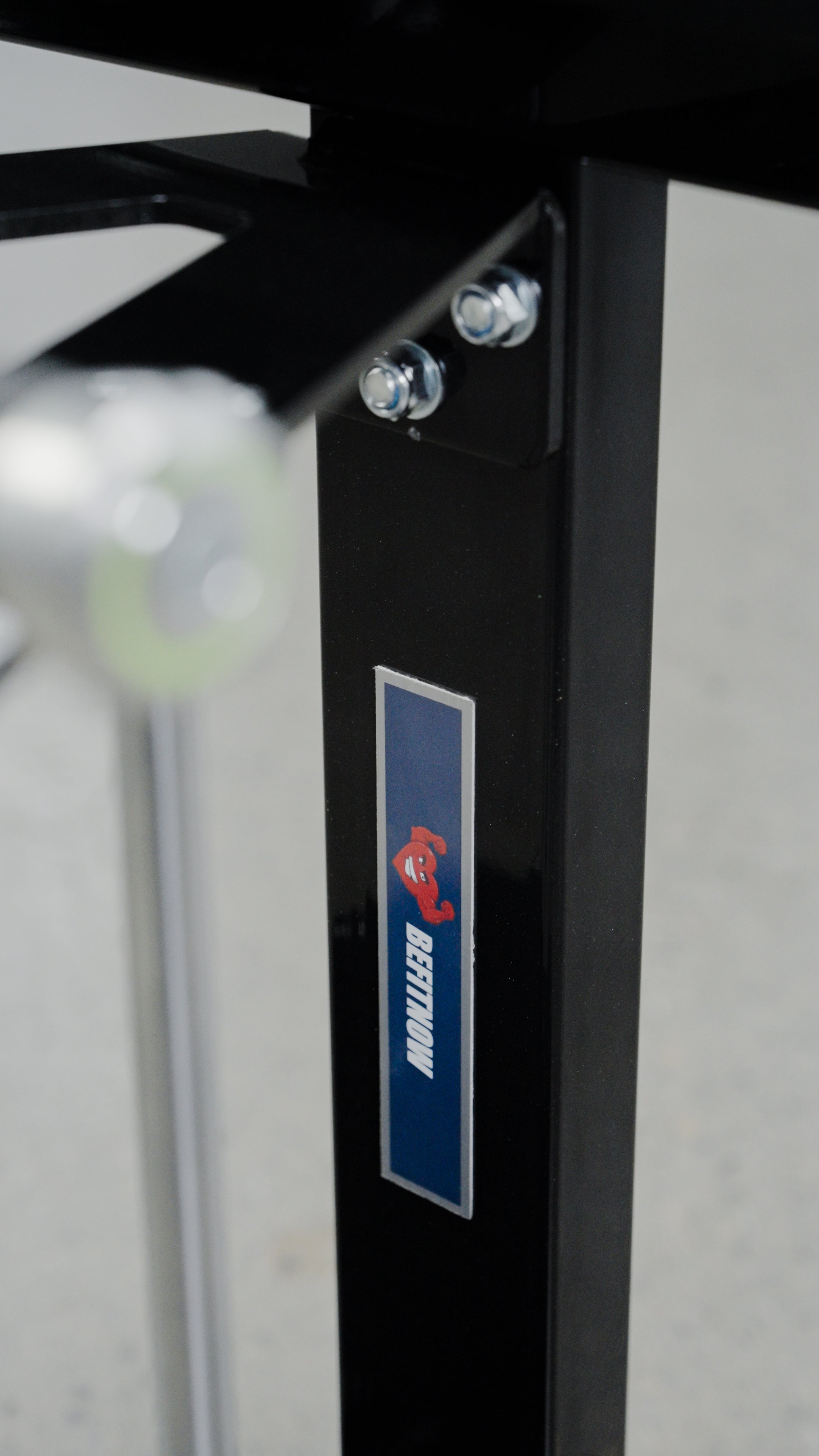 A close-up of the black metal frame from the affordable BefitNow Canada Forearm Trainer, featuring a blue BENCHON sticker and red logo. Four bolts secure a bracket to the frame, with the background softly blurred.