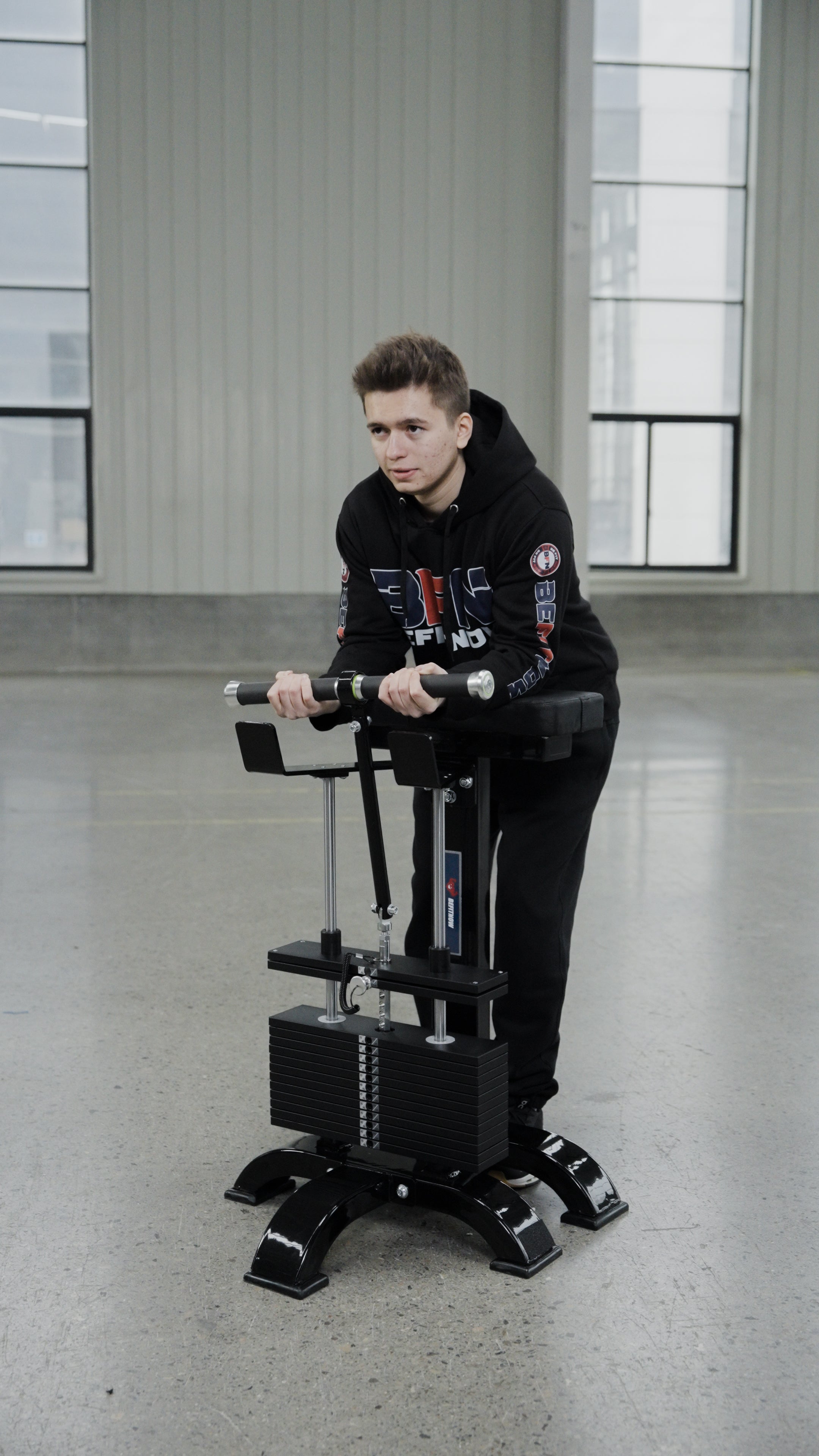 A person in a black hoodie uses the BefitNow Canada Forearm Trainer indoors, gripping its handlebars. The industrial setting features large windows, concrete floors, and gray walls, highlighting affordable forearm equipment.