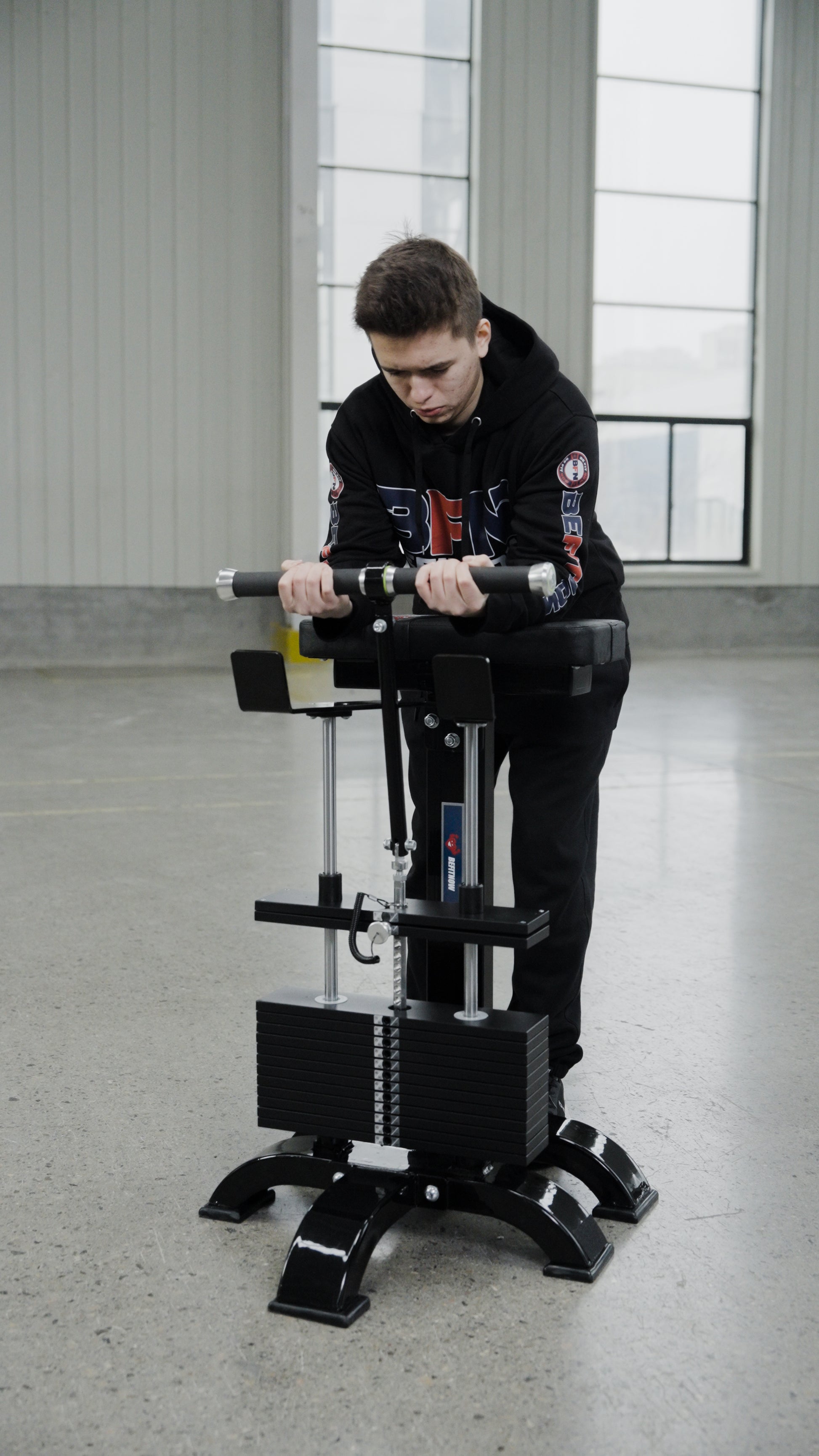 A person in a black hoodie is using the BefitNow Canada Forearm Trainer, an affordable fitness machine, in a spacious indoor area with large windows and a concrete floor.