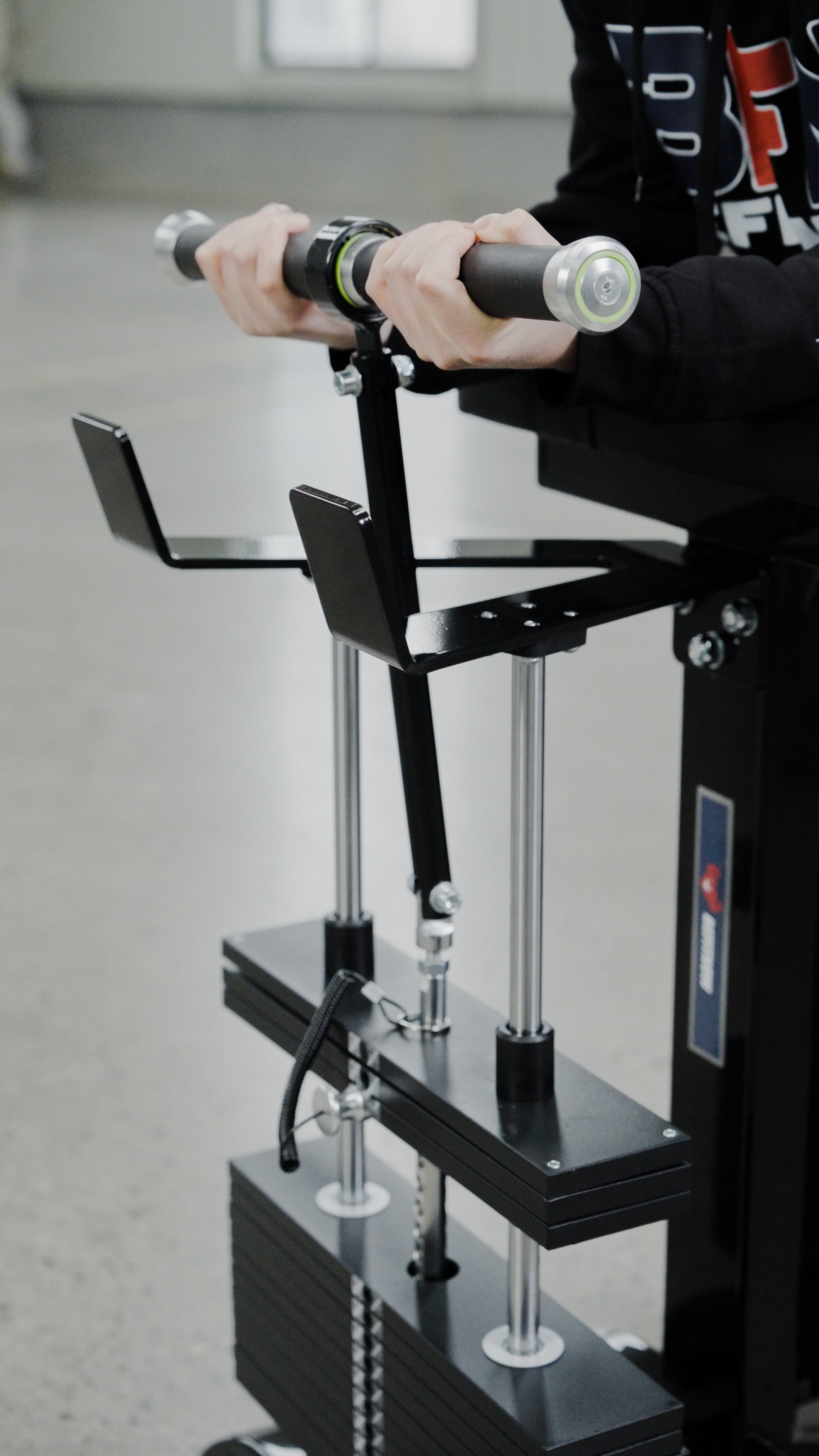 A person holds the handles of the BefitNow Canada Forearm Trainer, a black selectorized machine with adjustable weights and metal rods, set indoors on a smooth gray floor.