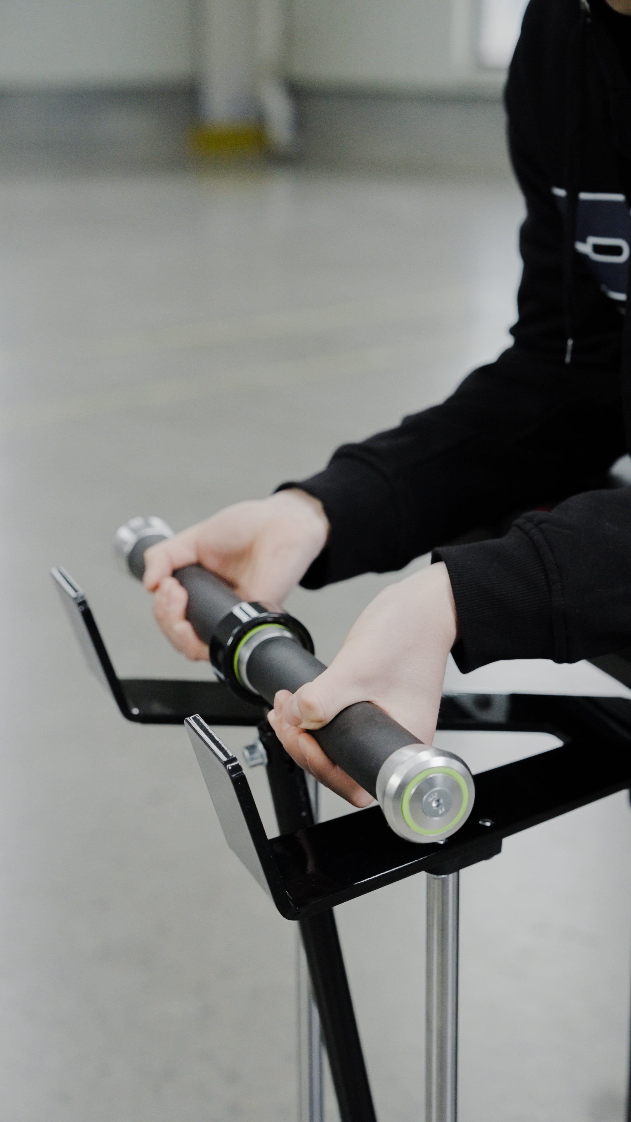 A person in a black hoodie grips the BefitNow Canada Forearm Trainer, featuring cylindrical metal handles with green accents, set against a blurred indoor background.