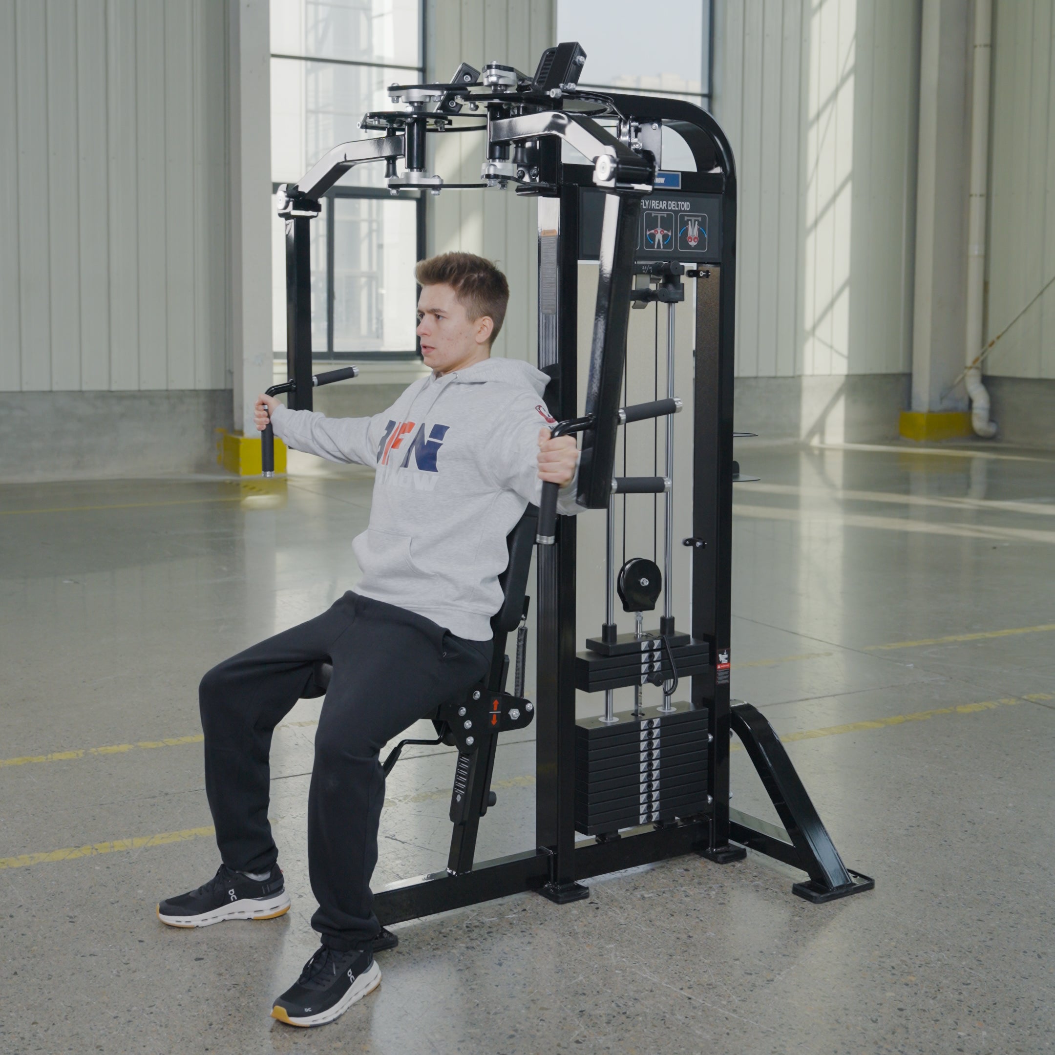 Wearing a gray hoodie and black pants, a person uses the Pearl Delt/Pec Fly by BefitNow Canada in a bright gym with large windows and concrete floors.