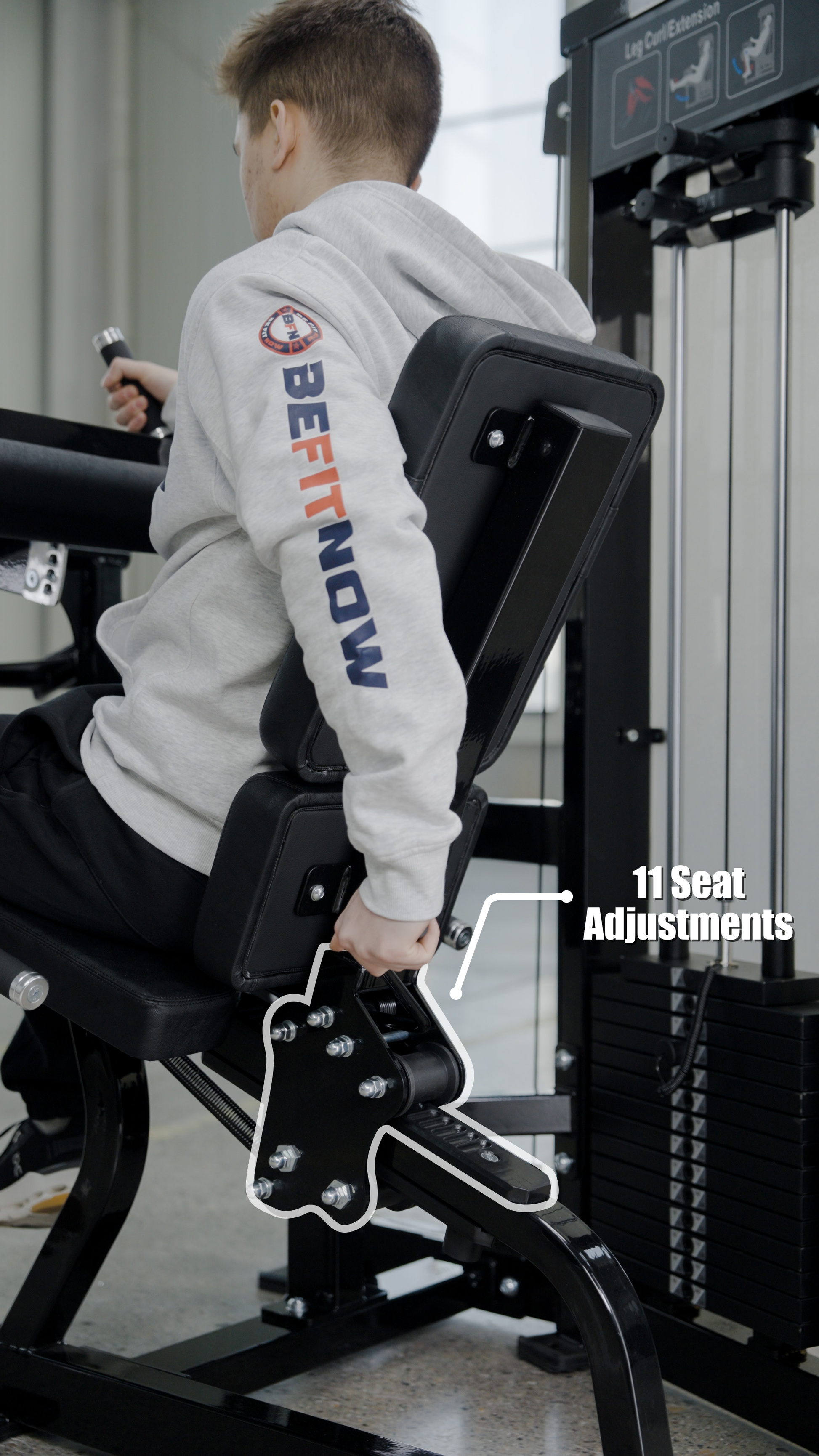 Wearing a gray hoodie, a person uses the BefitNow Canada Dual Function Leg Extension Leg Curl Machine at the gym, adjusting the lever with 11 seat adjustments. Heavy-duty equipment and weight stacks are visible in the background.
