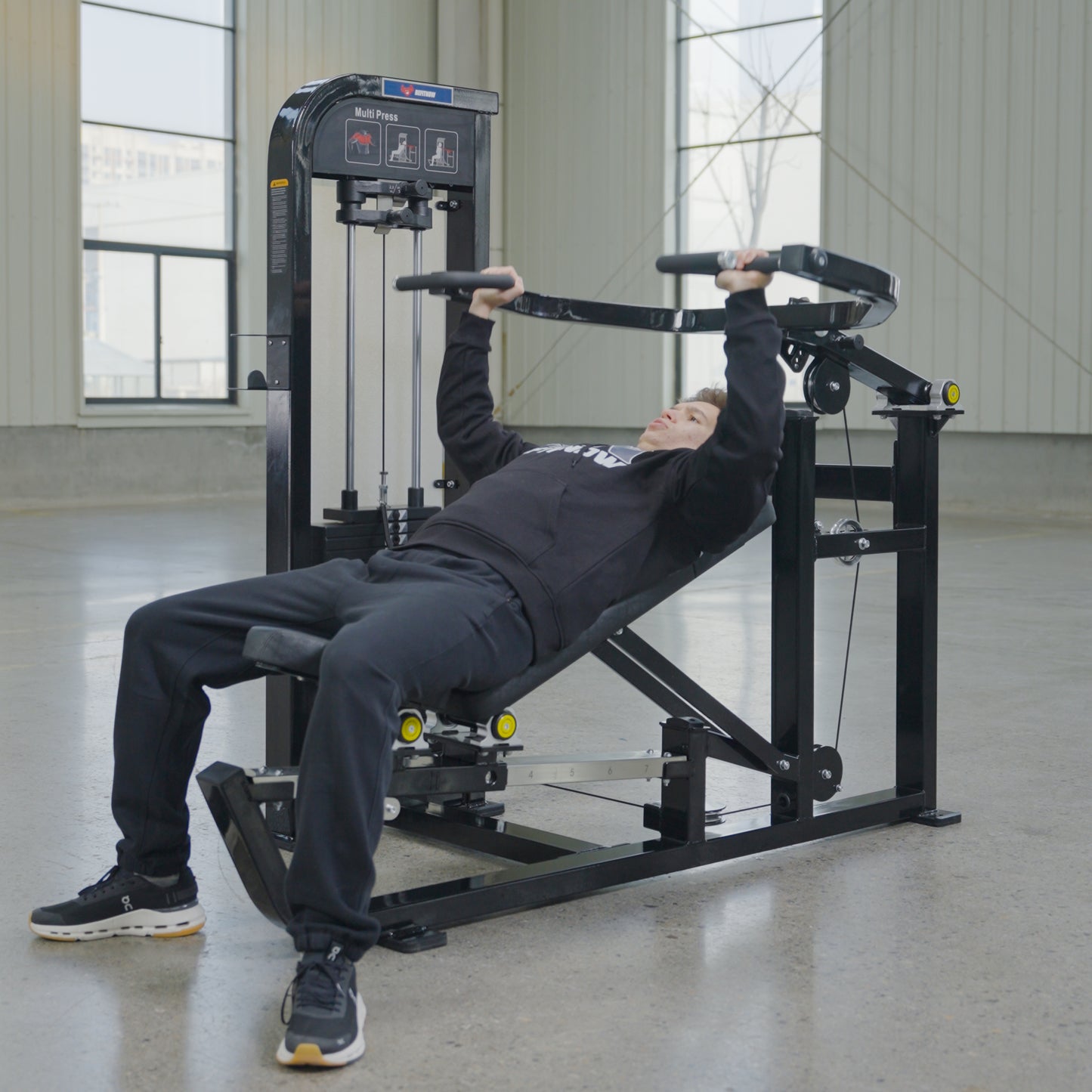 Wearing black athletic clothing, a person uses the BefitNow Canada Shoulder Press/Seated Chest Press with a 595lb weight stack in a spacious, well-lit gym with large windows and concrete floors.