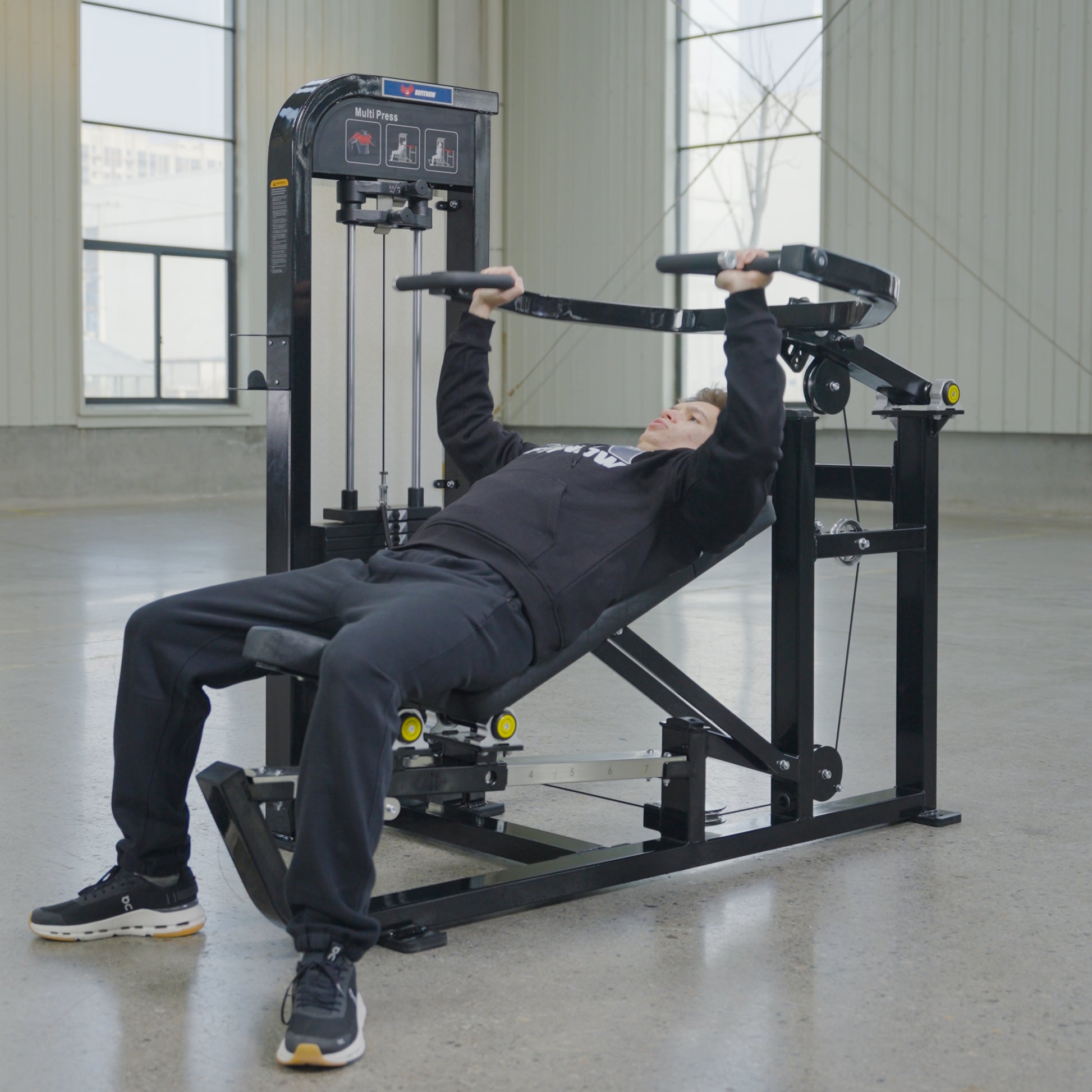 Wearing black athletic clothing, a person uses the BefitNow Canada Shoulder Press/Seated Chest Press with a 595lb weight stack in a spacious, well-lit gym with large windows and concrete floors.