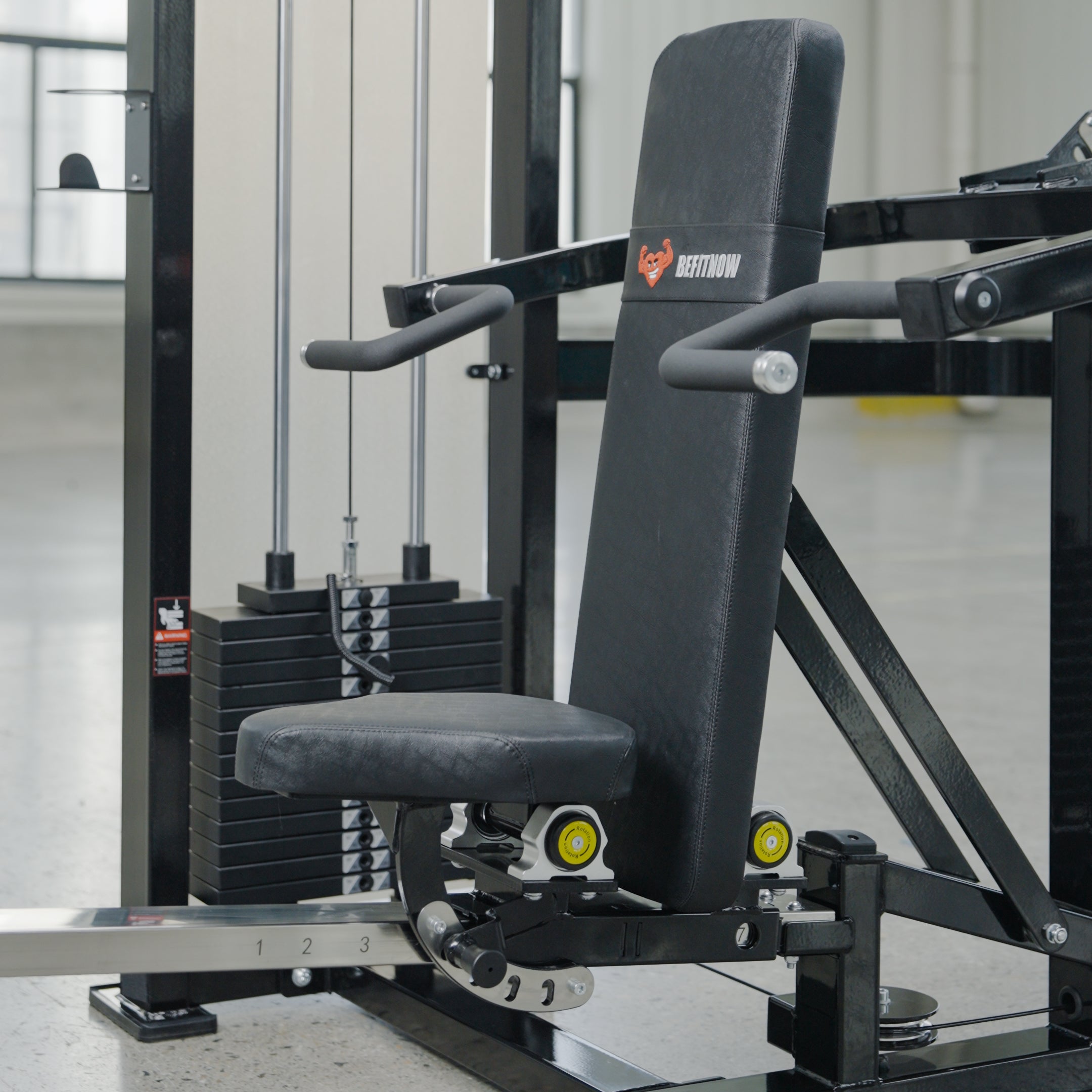 A close-up of the BefitNow Canada Shoulder Press/Seated Chest Press shows its black adjustable design with padded seat/backrest, weight stack, and metal arms with handles, positioned on a light-colored gym floor.