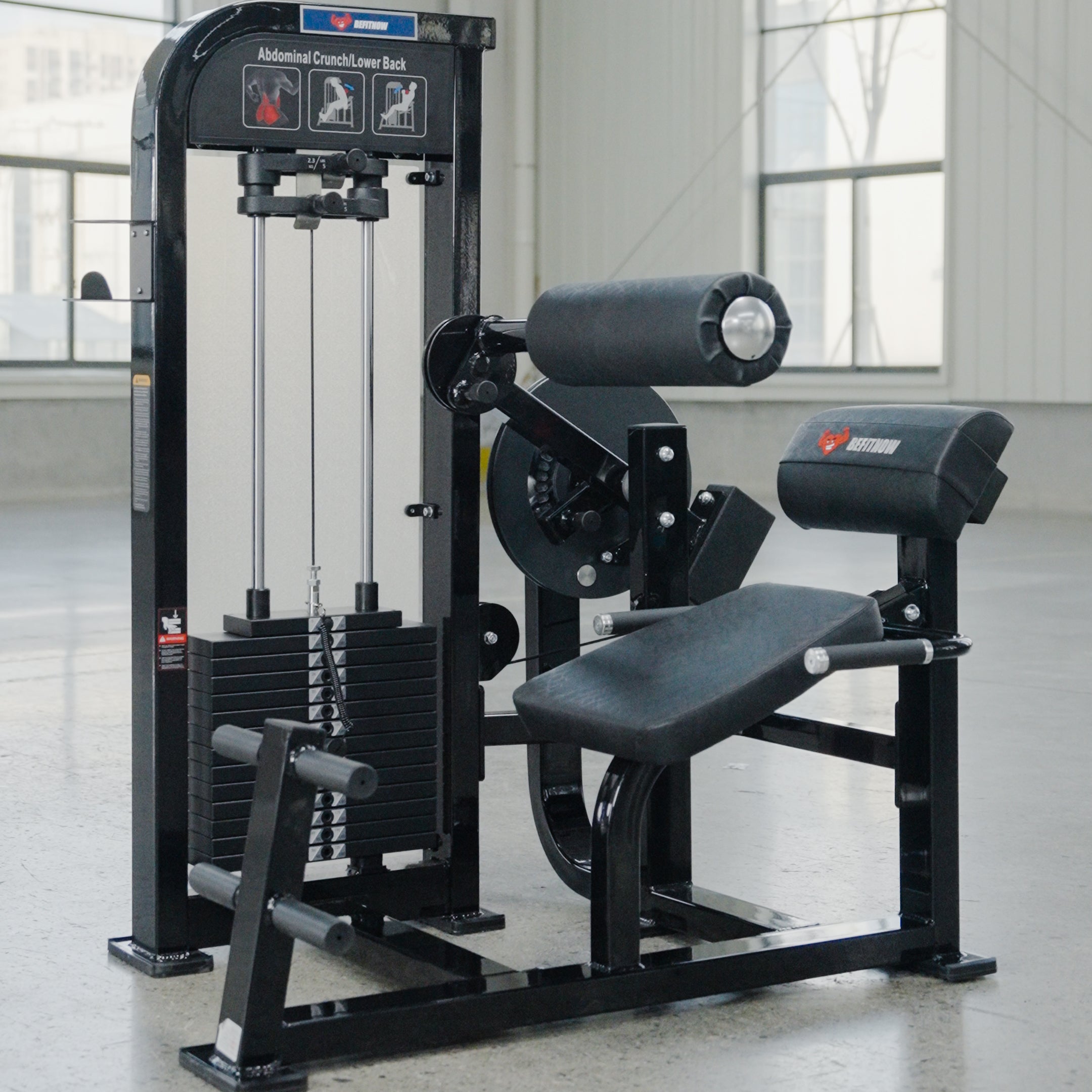 The BefitNow Canada Abdominal Crunch/Low Back is a black machine for core and lower back workouts, featuring padded rollers, a seat, weight stack, and handles; pictured in a bright room with large windows.