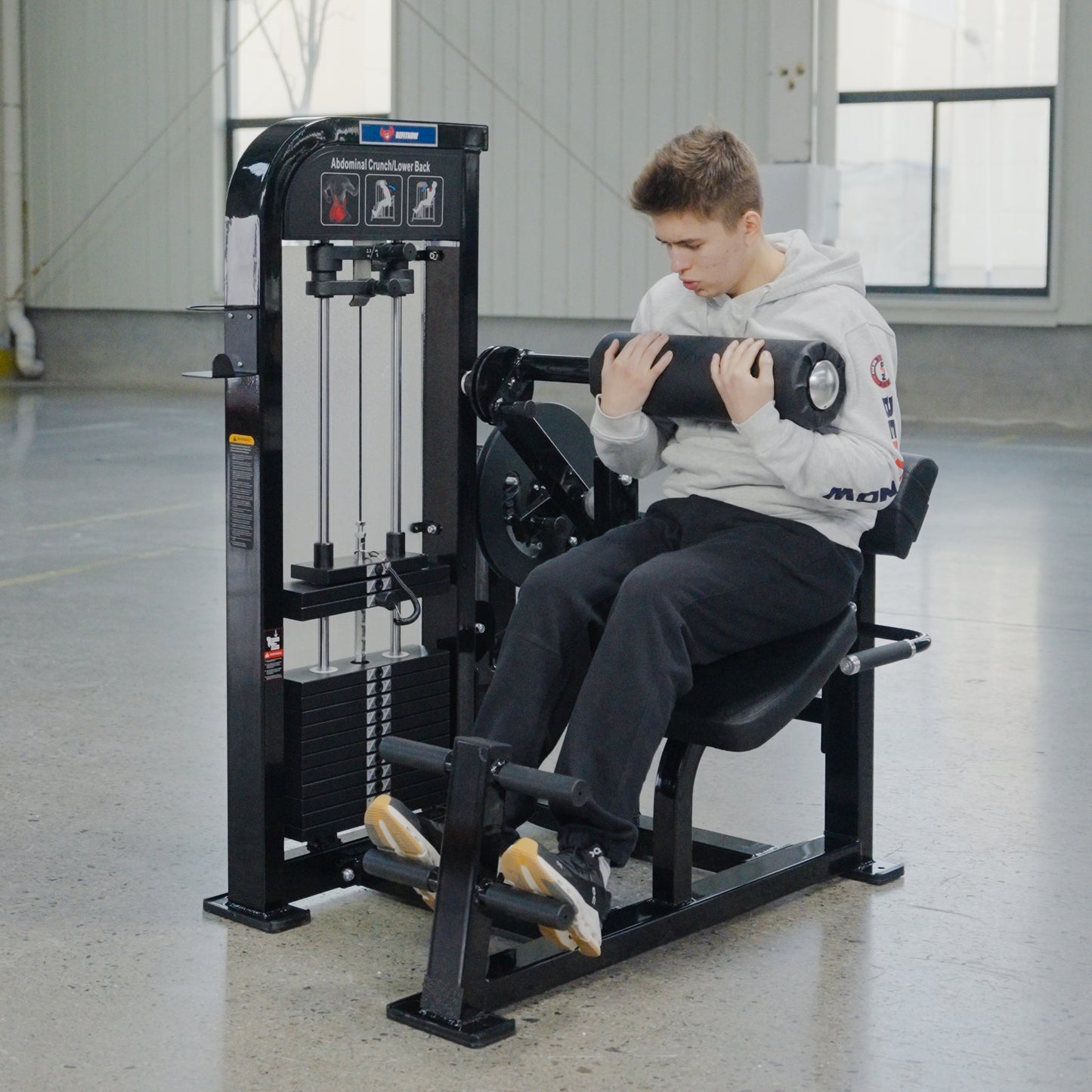 A person in a light gray hoodie and dark pants uses the BefitNow Canada Abdominal Crunch/Low Back machine in a bright, spacious gym with large windows and polished concrete floors, focusing on core strength.