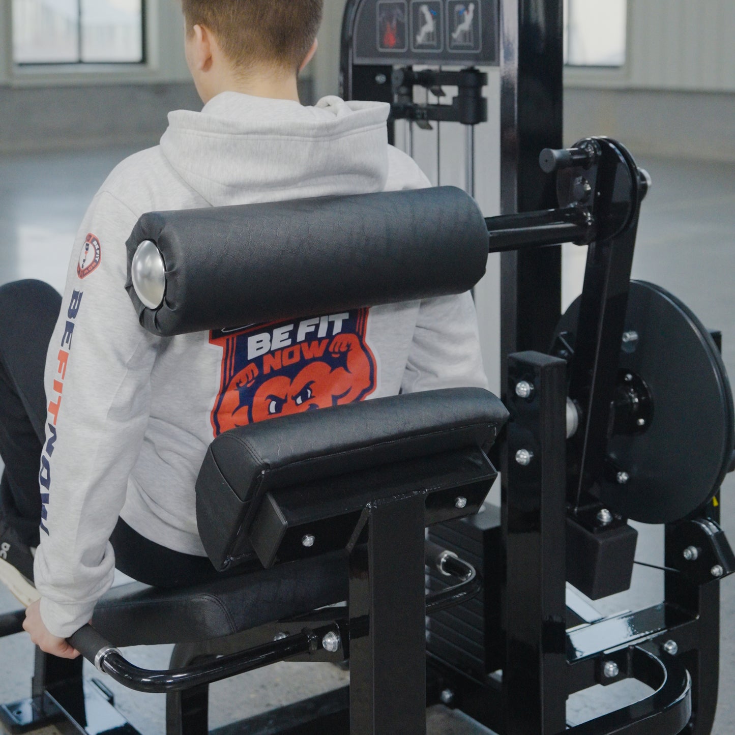 Wearing a gray BE FIT NOW hoodie, a person uses a seated leg extension machine in a gym with large windows, fitness equipment, and a BefitNow Canada Abdominal Crunch/Low Back machine for core workouts.