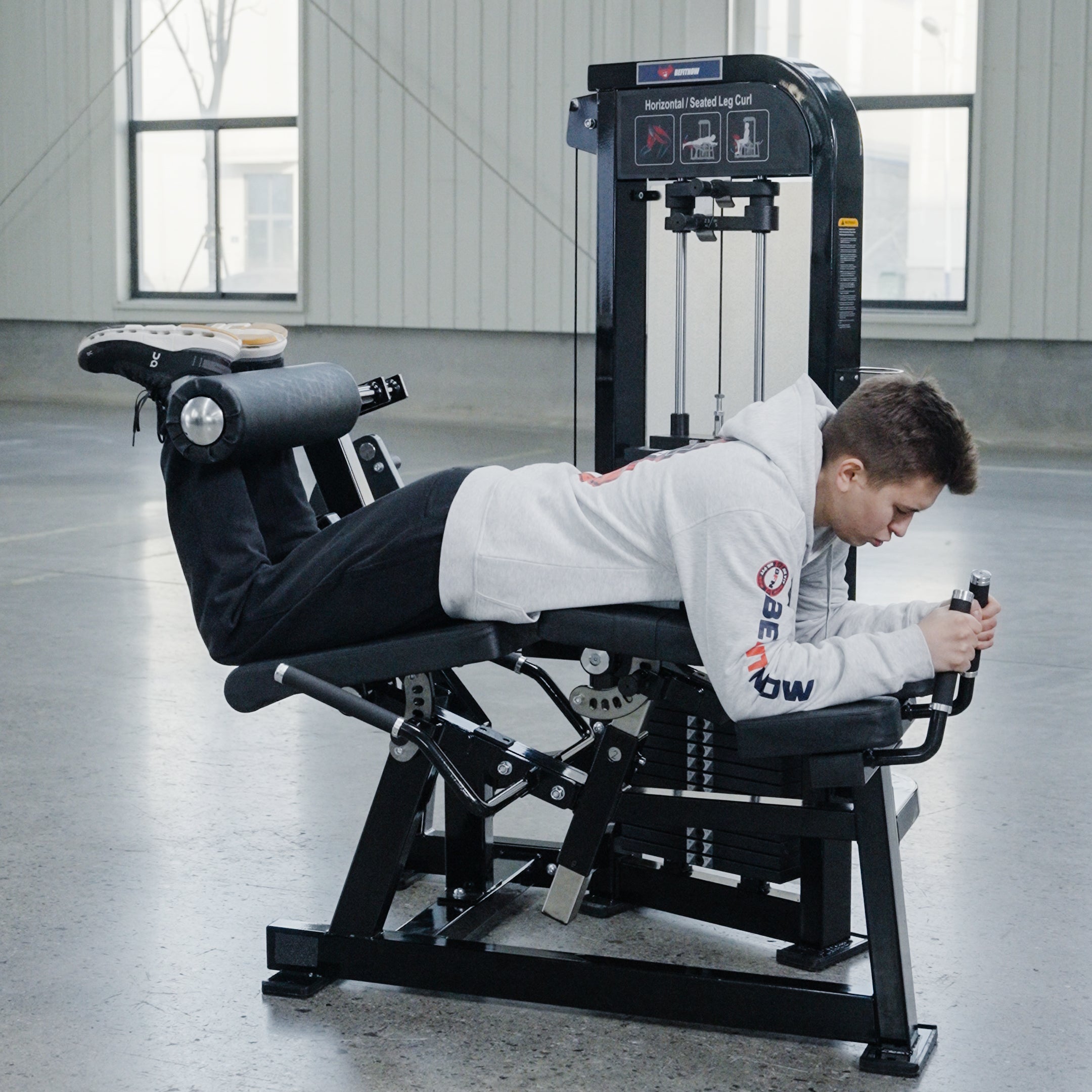 A person in a light hoodie and dark pants uses the BefitNow Canada Leg Extension/Lying Down Leg Curl, gripping the handles and lifting the padded bar in a gym with large windows and commercial equipment.