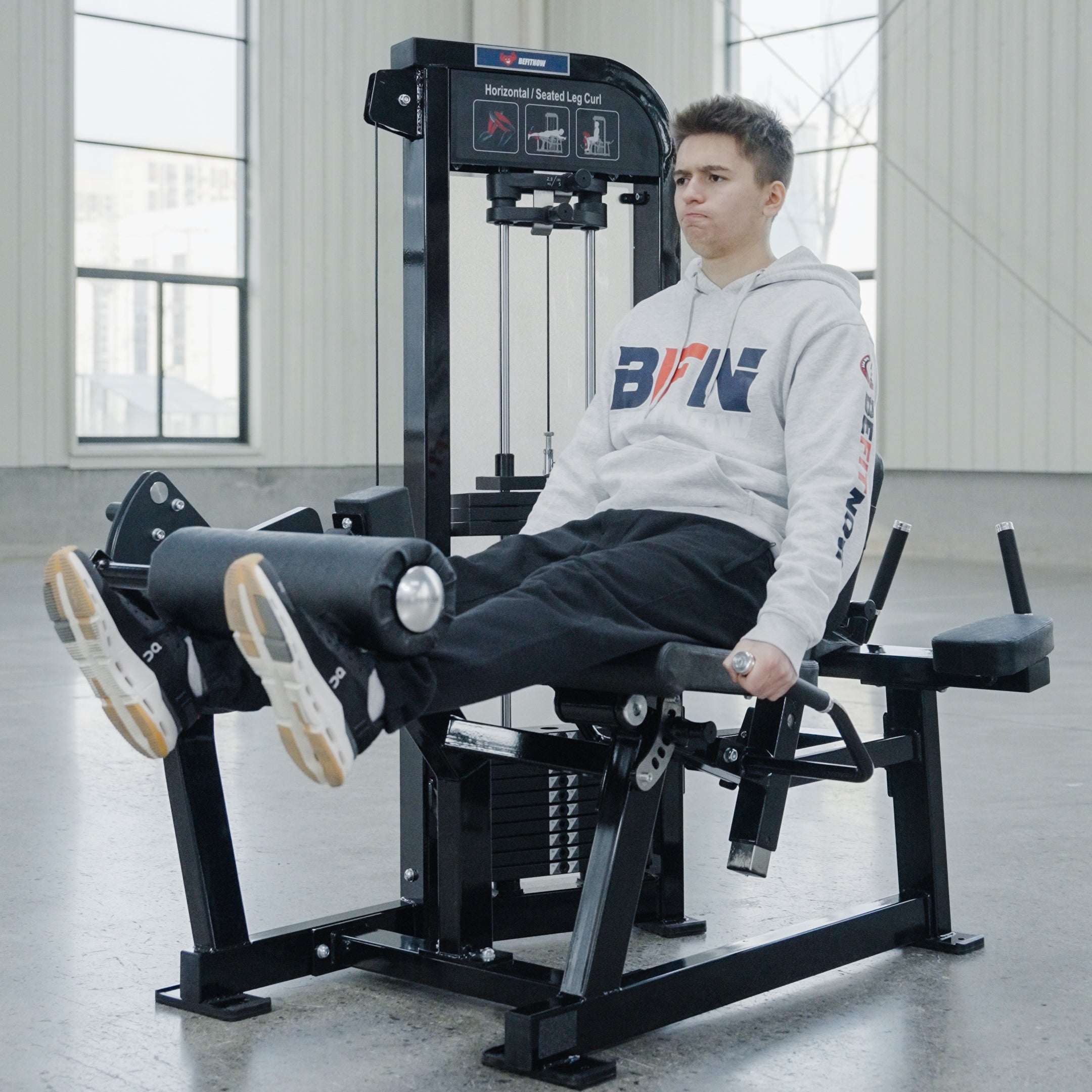A young man wearing a light gray hoodie and black pants uses the BefitNow Canada Leg Extension/Lying Down Leg Curl machine in a bright, spacious gym, focusing intently as he works out.