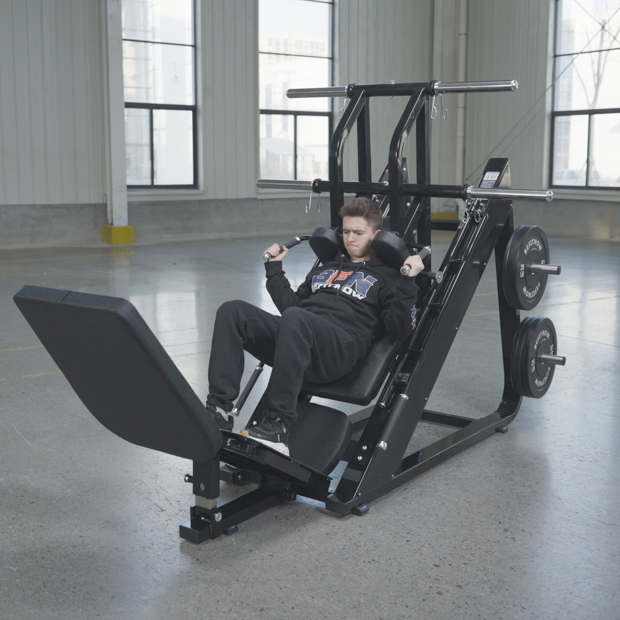 A person in athletic clothing uses the BefitNow Canada 45 Degree Leg Press Hack Squat—measuring 93"x65"x57" and weighing 560 lbs—in a spacious gym with large windows and concrete floors, pushing the platform with weight plates loaded.