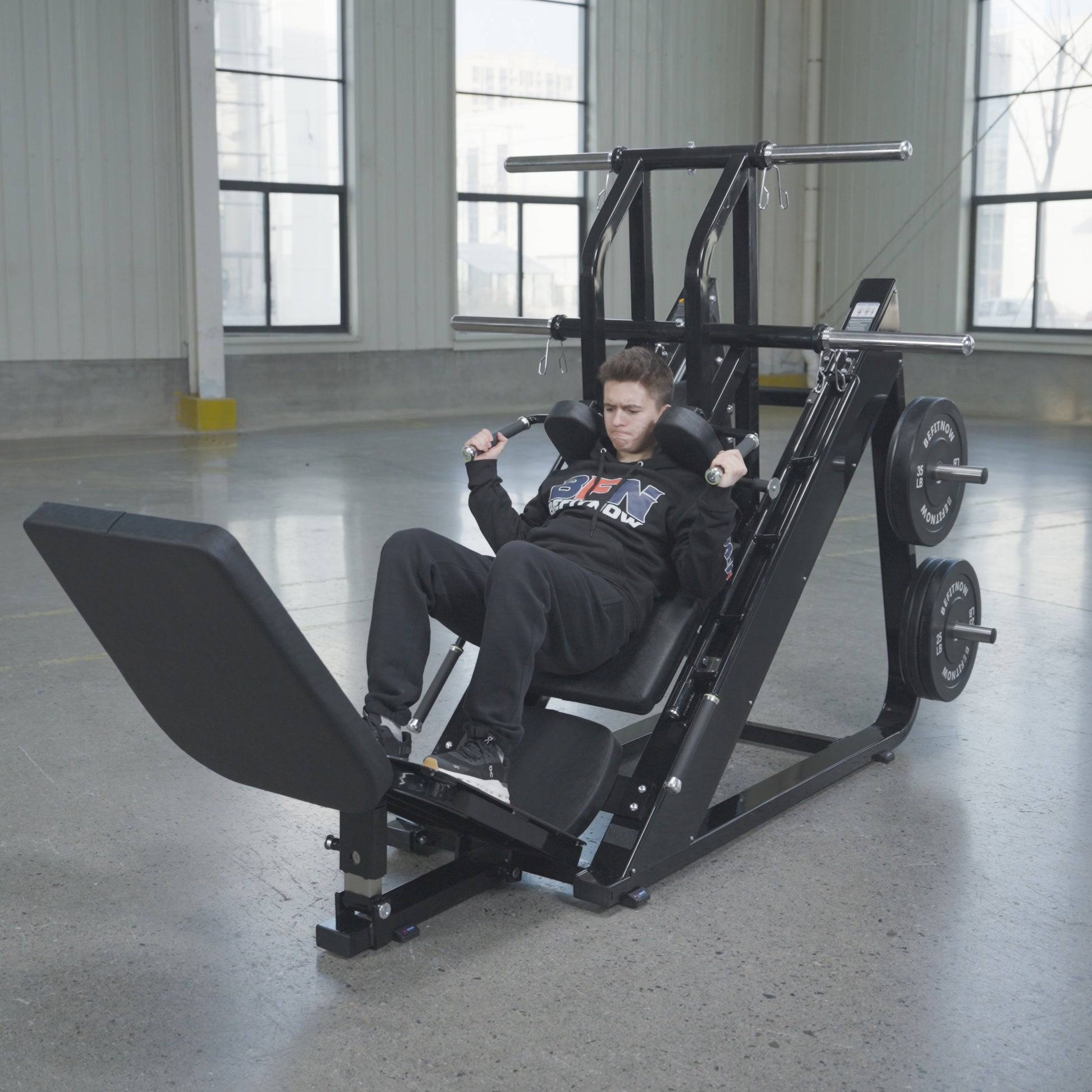 A person in athletic clothing uses the BefitNow Canada 45 Degree Leg Press Hack Squat—measuring 93"x65"x57" and weighing 560 lbs—in a spacious gym with large windows and concrete floors, pushing the platform with weight plates loaded.