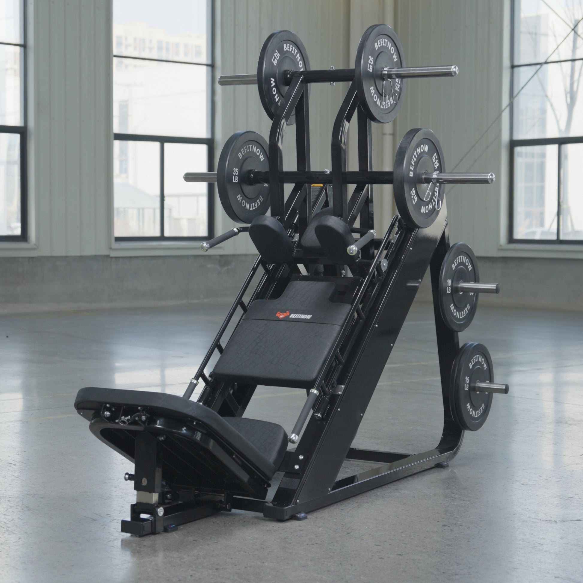 The BefitNow Canada 45 Degree Leg Press Hack Squat, shown in black and loaded with 560lbs of weight plates, sits in a spacious gym with large windows and concrete floors. Dimensions: 93"x65"x57".