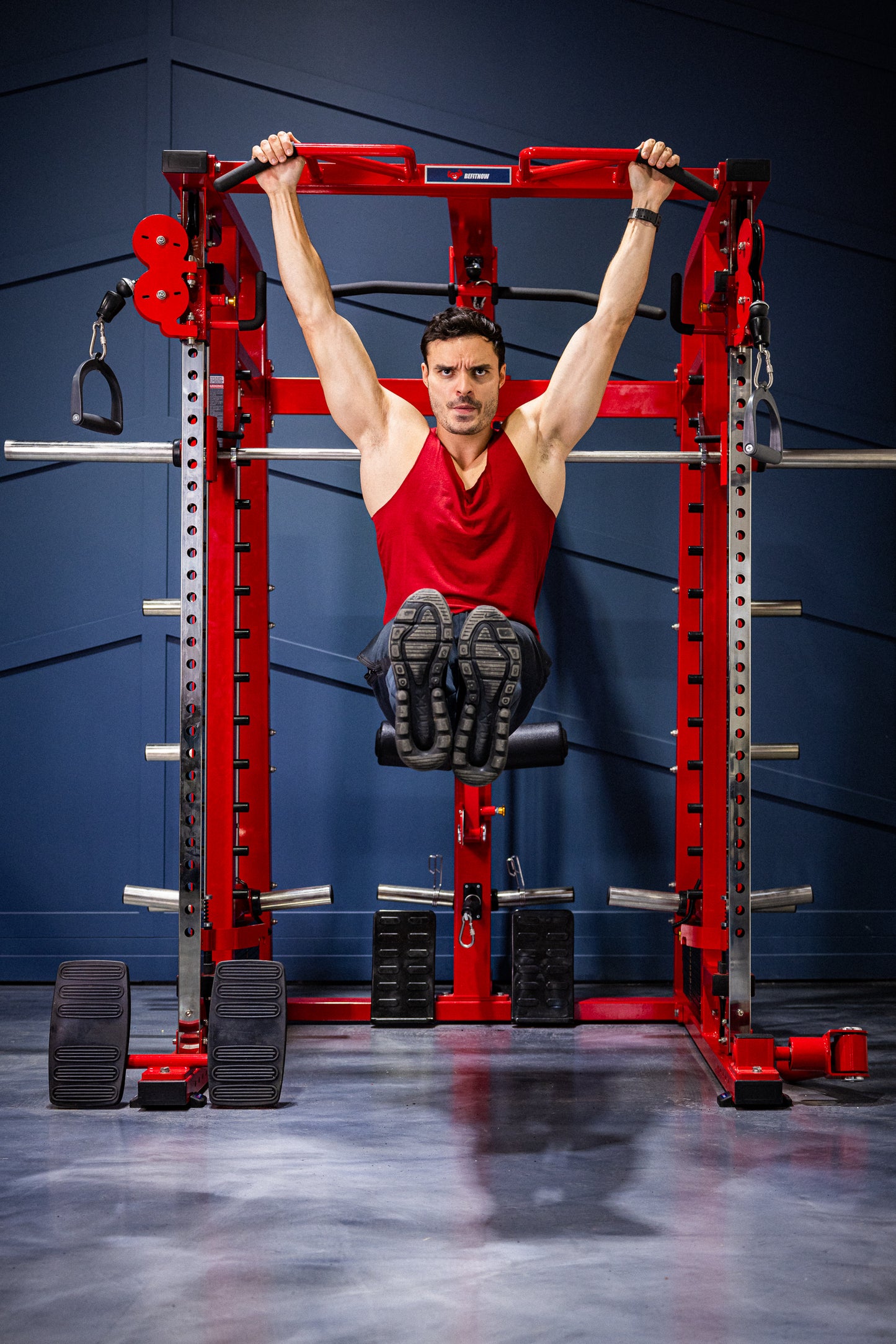 A man in a red tank top performs a hanging leg raise on the BefitNow Canada Mr. Monster Smith Machine Functional Trainer, gripping the overhead bar of the red rack in a gym as he lifts his legs straight out.