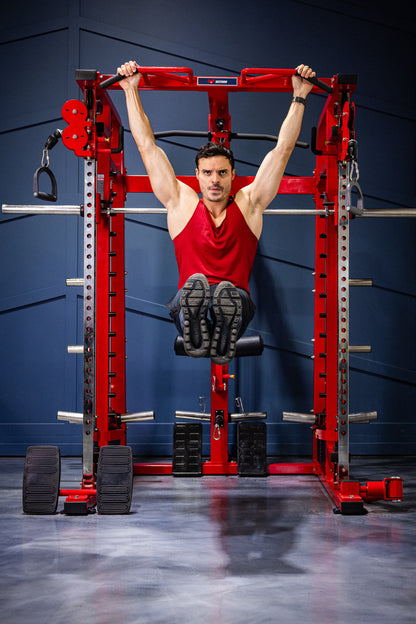 A man in a red tank top performs a hanging leg raise on the BefitNow Canada Mr. Monster Smith Machine Functional Trainer, gripping the overhead bar of the red rack in a gym as he lifts his legs straight out.