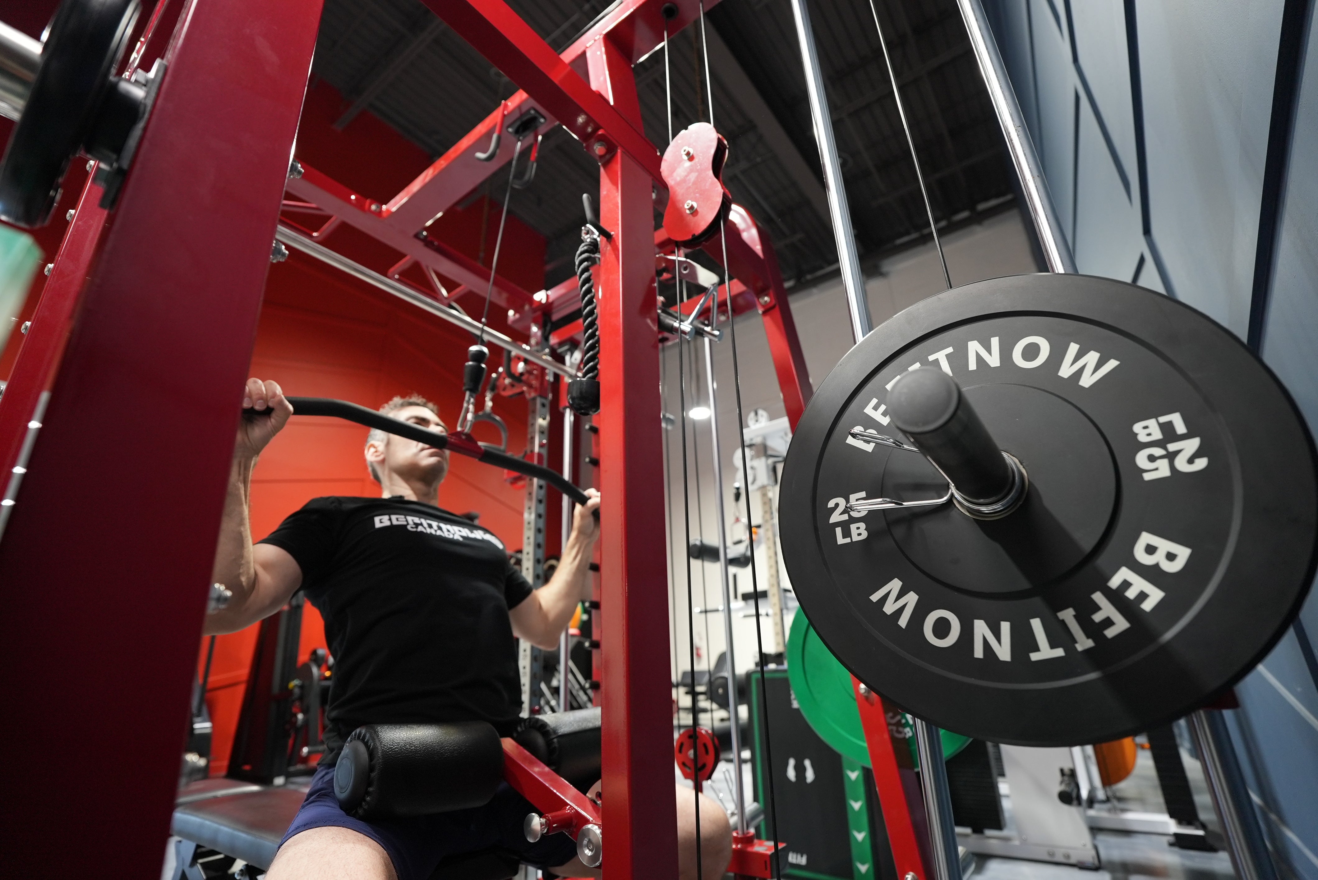 A person in a black shirt uses the BefitNow Canada Mr. Monster Smith Machine Functional Trainer at the gym, gripping the bar while seated. A 25 LB weight plate is visible on the machine in the foreground.