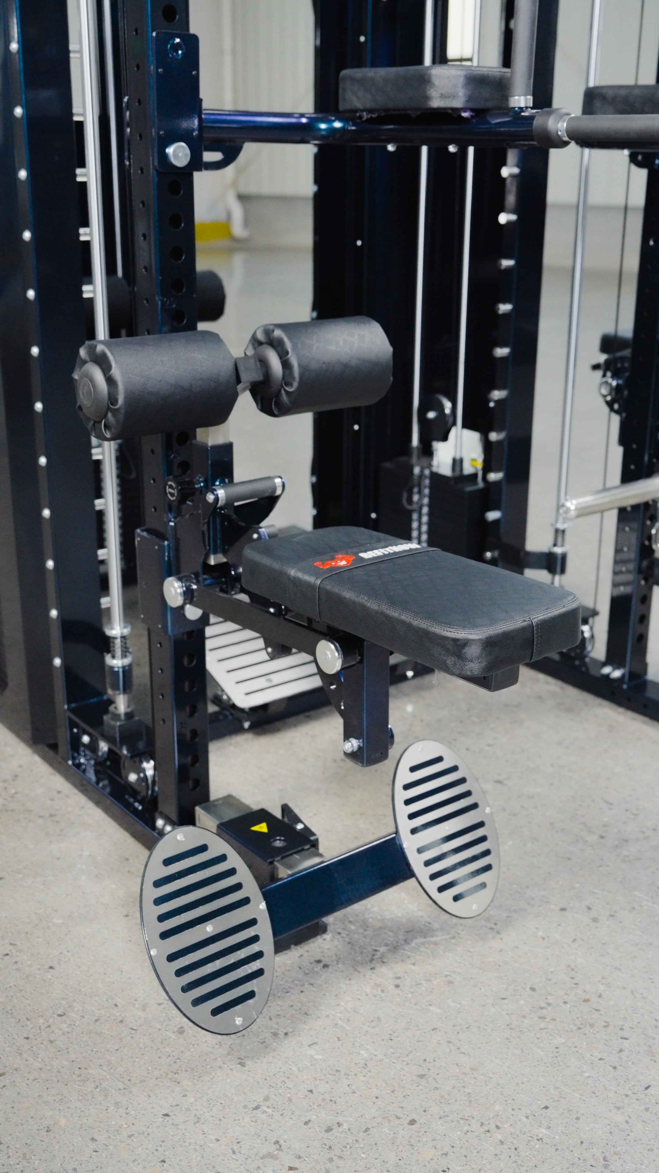 A close-up of BefitNow Canada's The Professor: The Kahuna on Steroids gym machine, showcasing padded rollers, a cushioned bench seat, and dual metal footplates for leg exercises on the fitness facility's concrete floor.