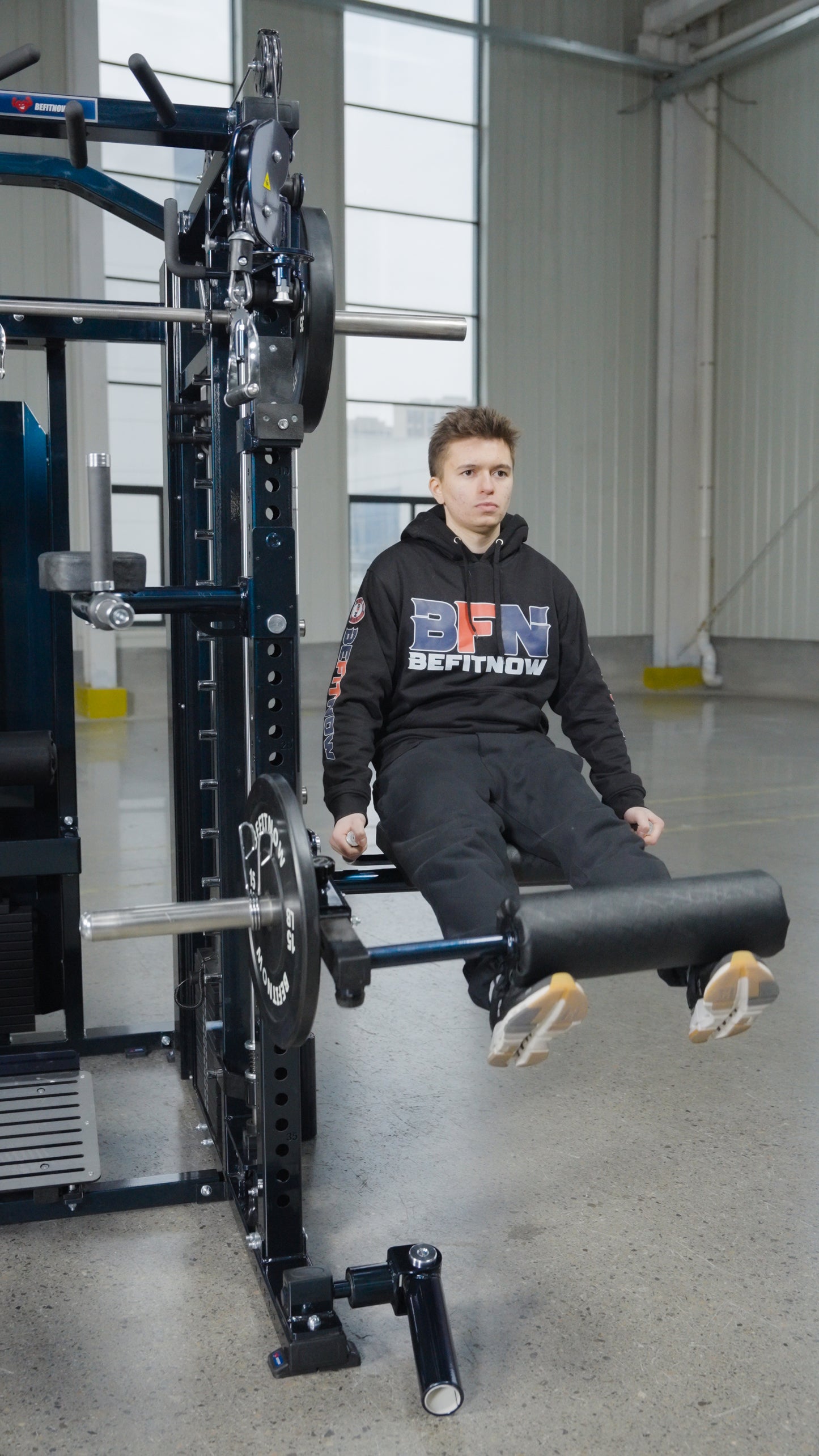 A person in a black hoodie and track pants uses The Professor by BefitNow Canada for leg extension exercises in a spacious indoor gym.